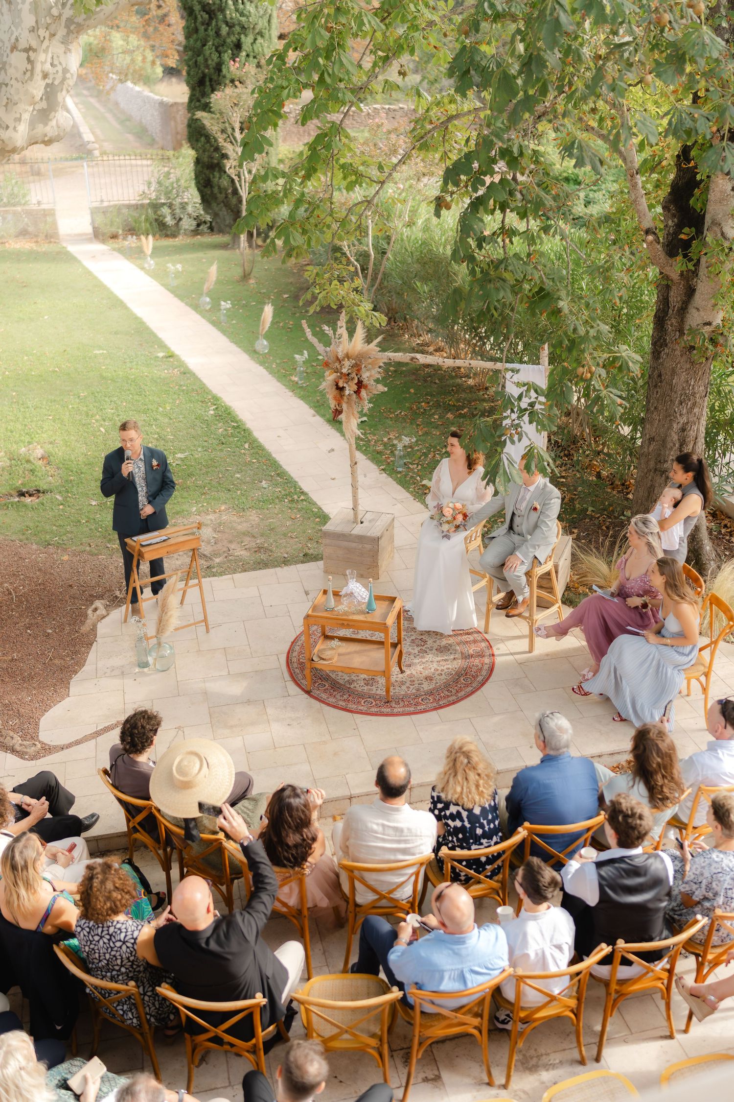 Aerial view of an outdoor wedding ceremony with guests seated on wooden chairs around a couple exchanging vows in a garden.