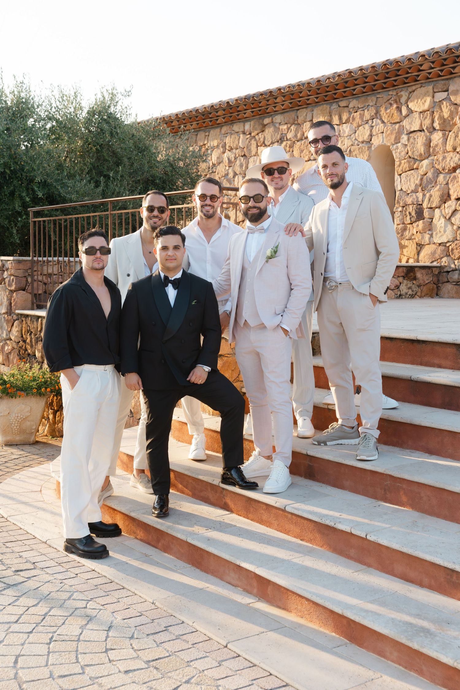 Groom with groomsmen in matching beige suits posing on stone steps at golden hour wedding venue.