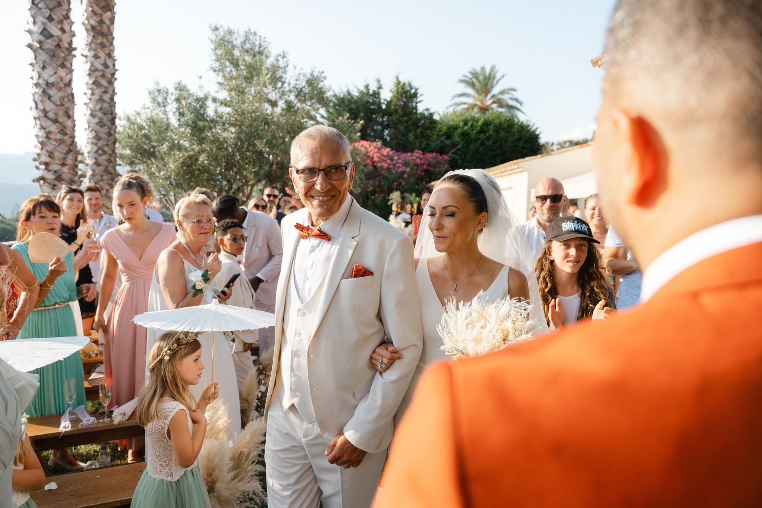 Bride walks down the aisle with an escort as smiling guests celebrate at a sunny outdoor wedding.