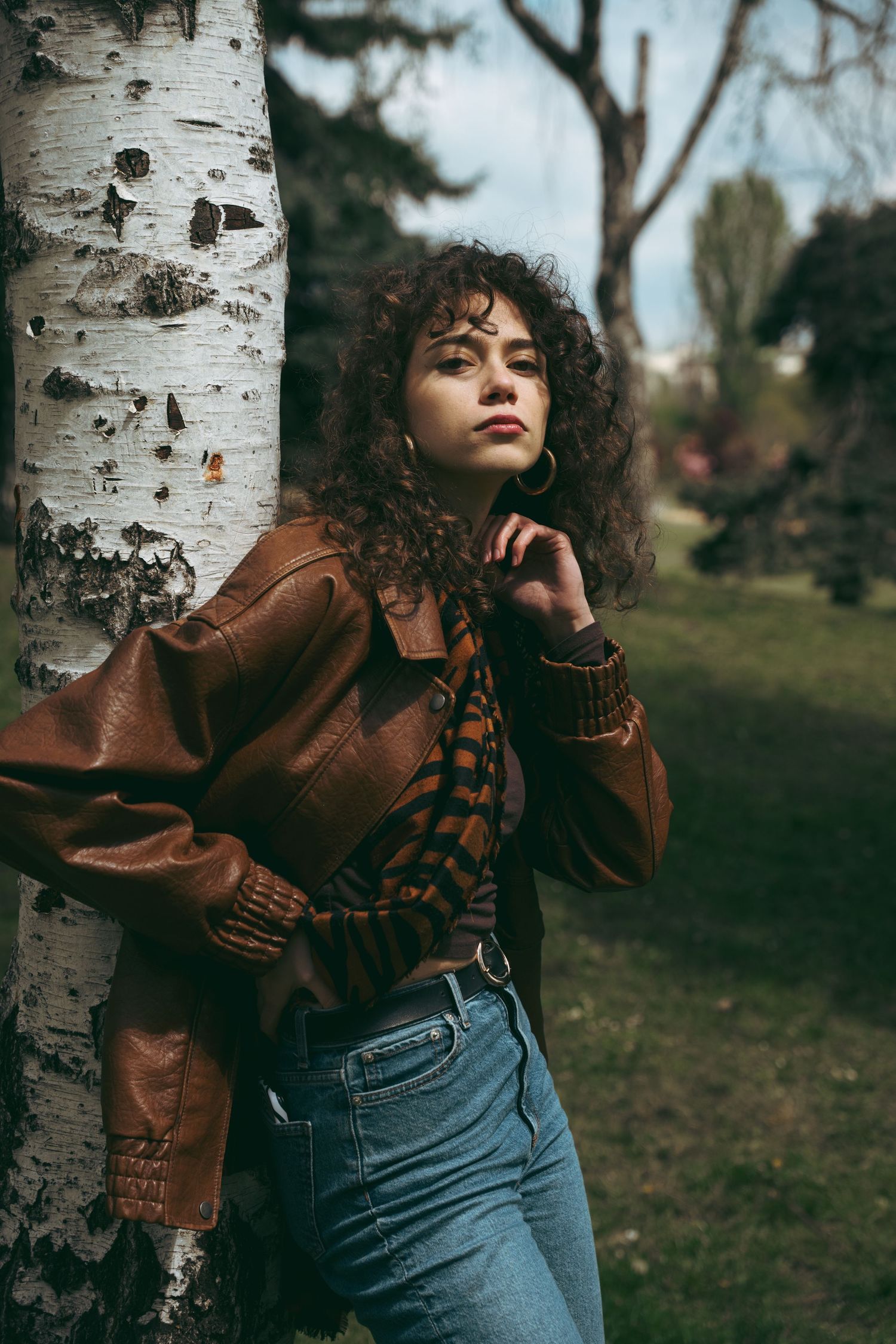 Young woman with curly hair in brown leather jacket leaning against birch tree in autumn park.