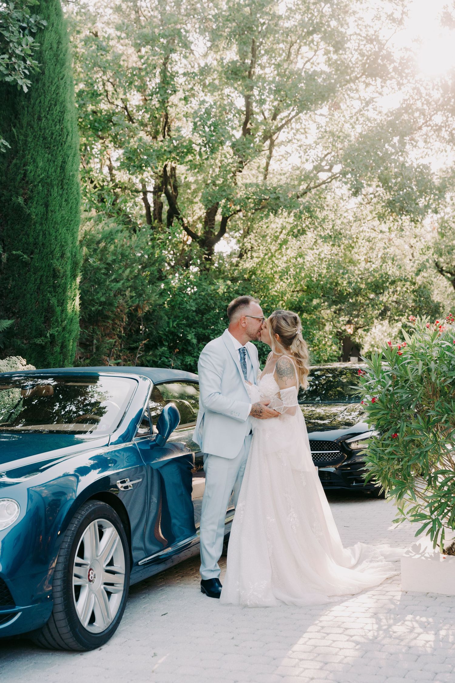 Bride and groom sharing a kiss beside a blue Bentley convertible in a lush garden.