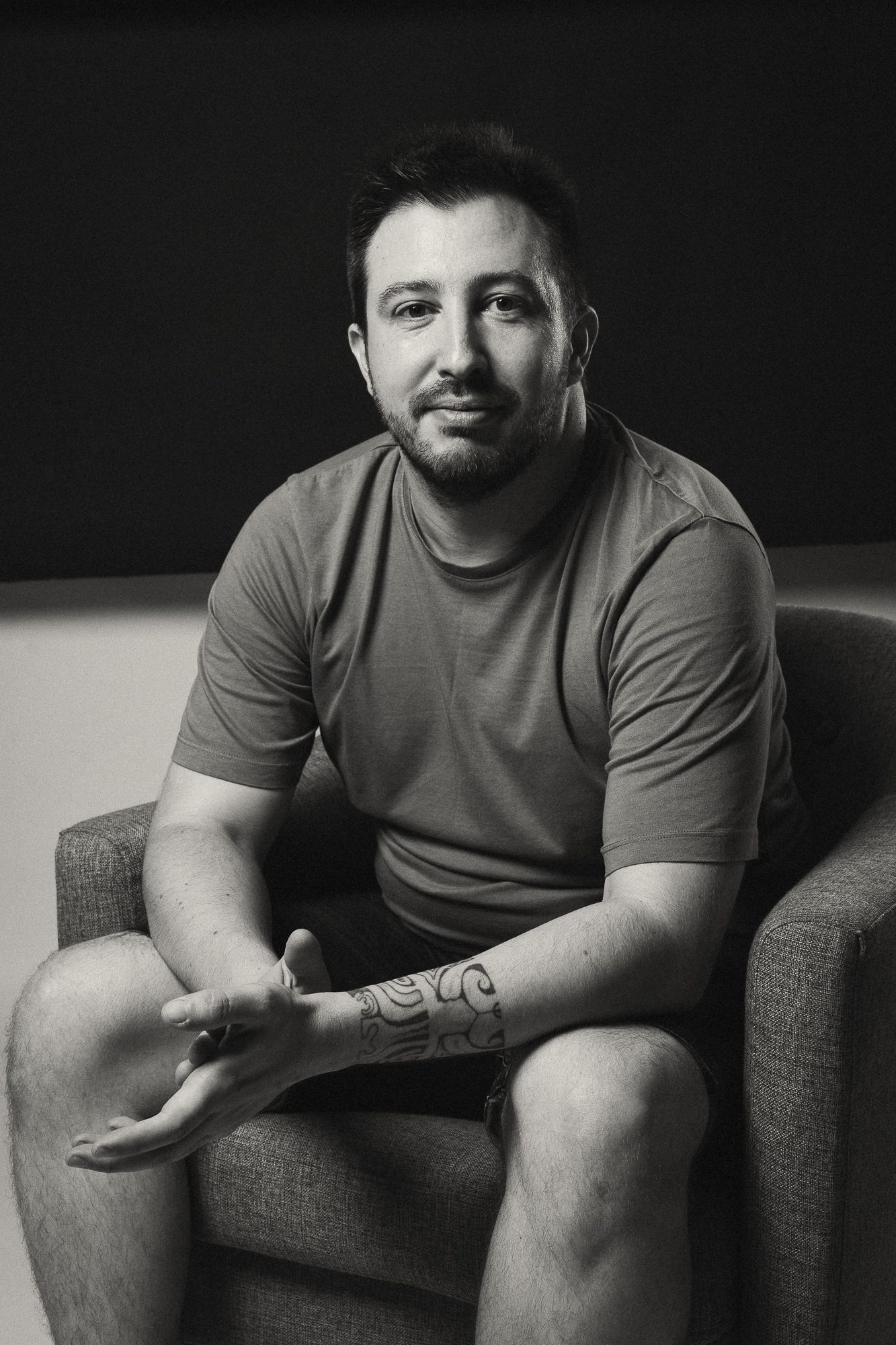Black and white portrait of tattooed man sitting relaxed in armchair looking at camera.