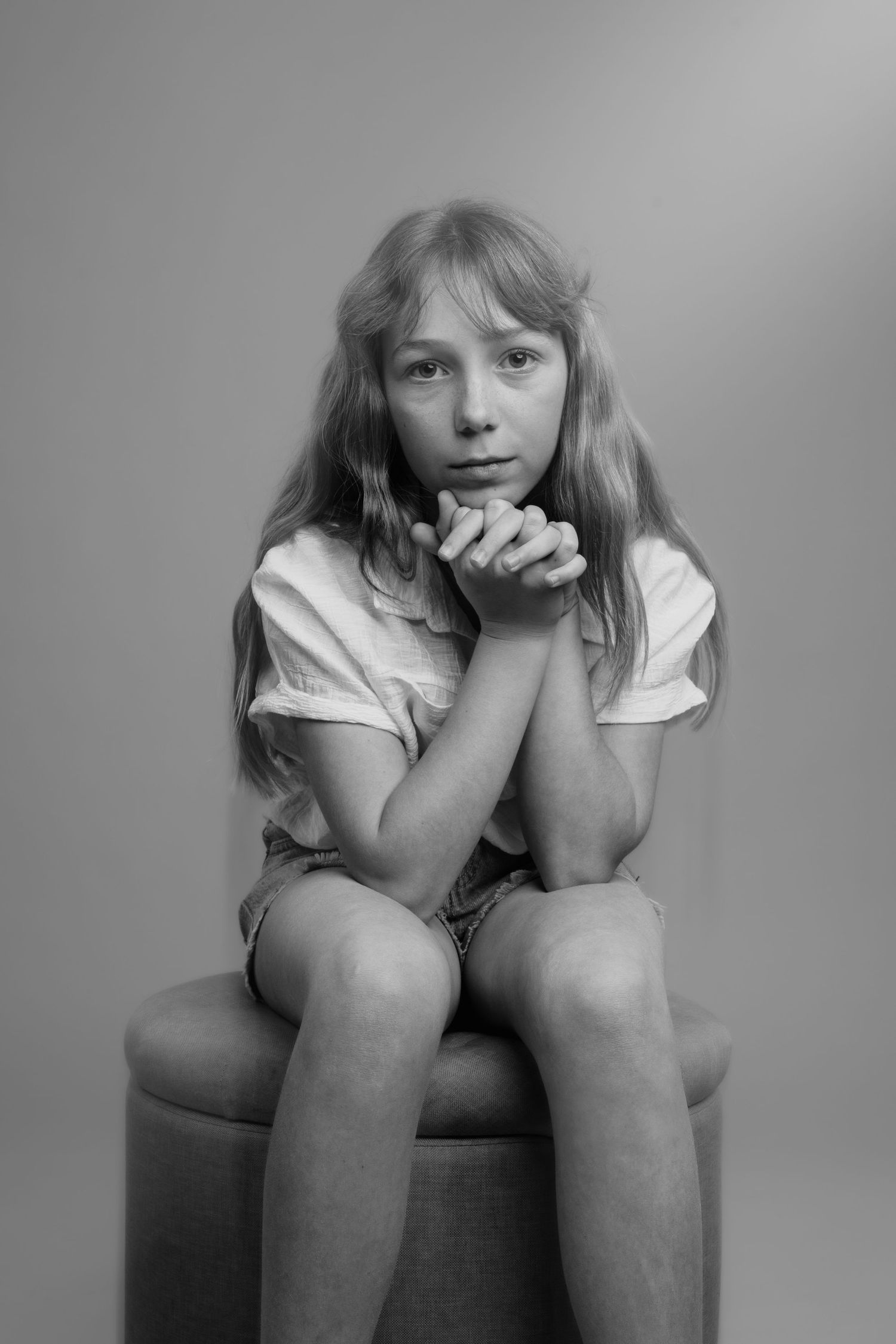 Black and white portrait of a young girl sitting on a stool with hands clasped under her chin.
