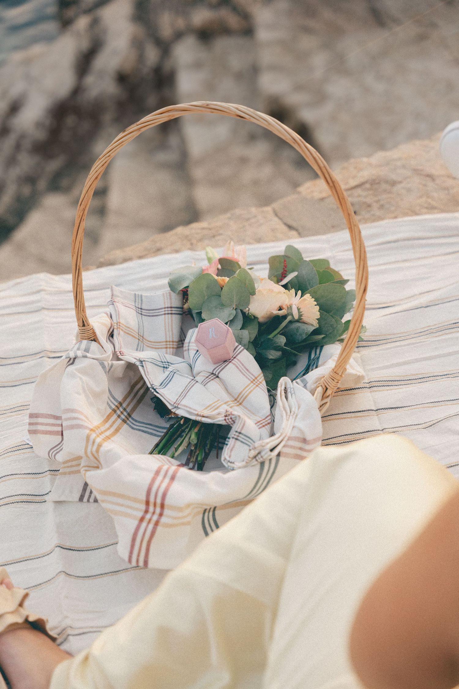 Wicker picnic basket with fresh flowers and plaid napkin on striped blanket for romantic outdoor celebration.