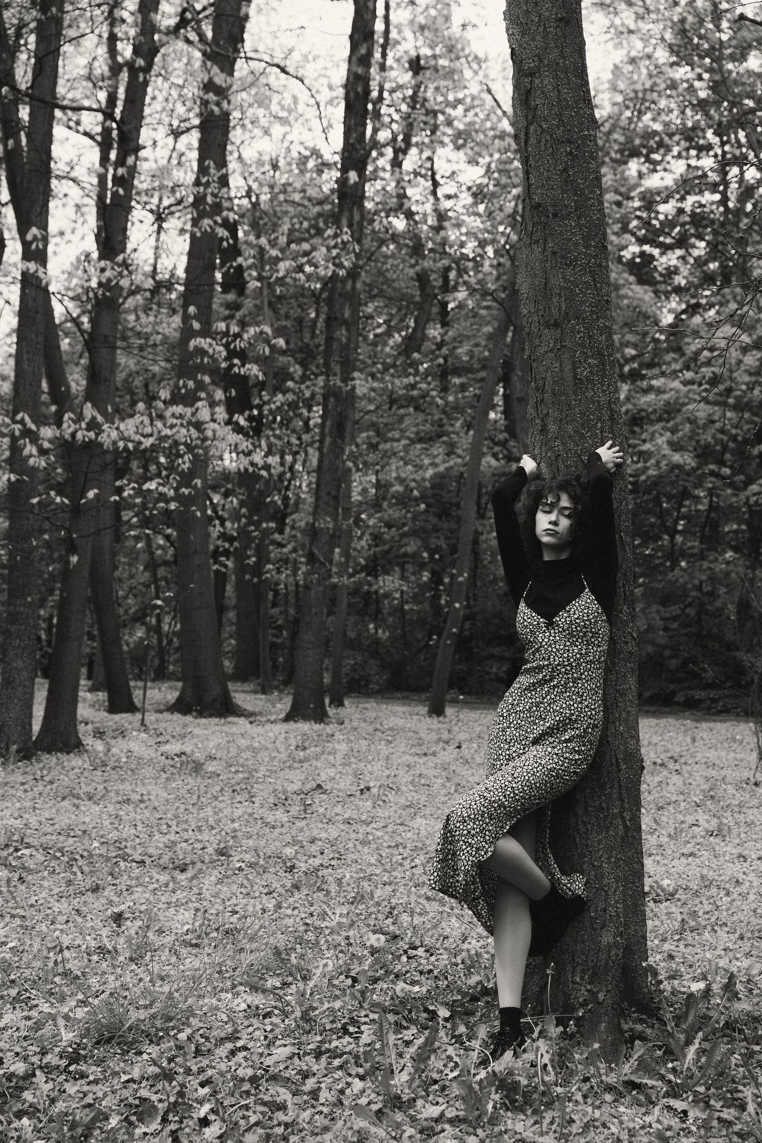 Black and white photo of curly-haired woman in floral dress posing against tree in forest.