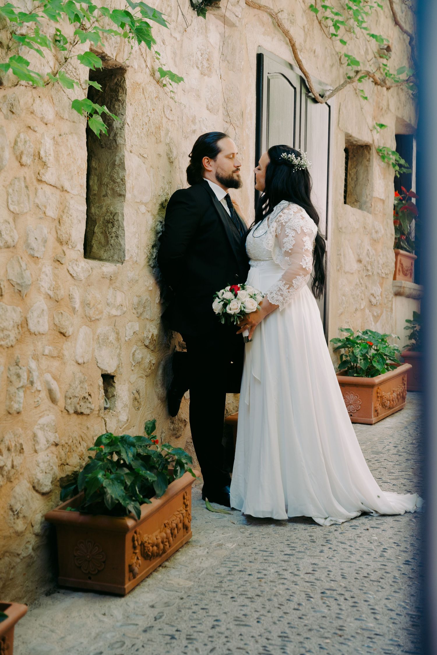 Bride and groom share a romantic moment outside a rustic stone building, bride holding a white bouquet.