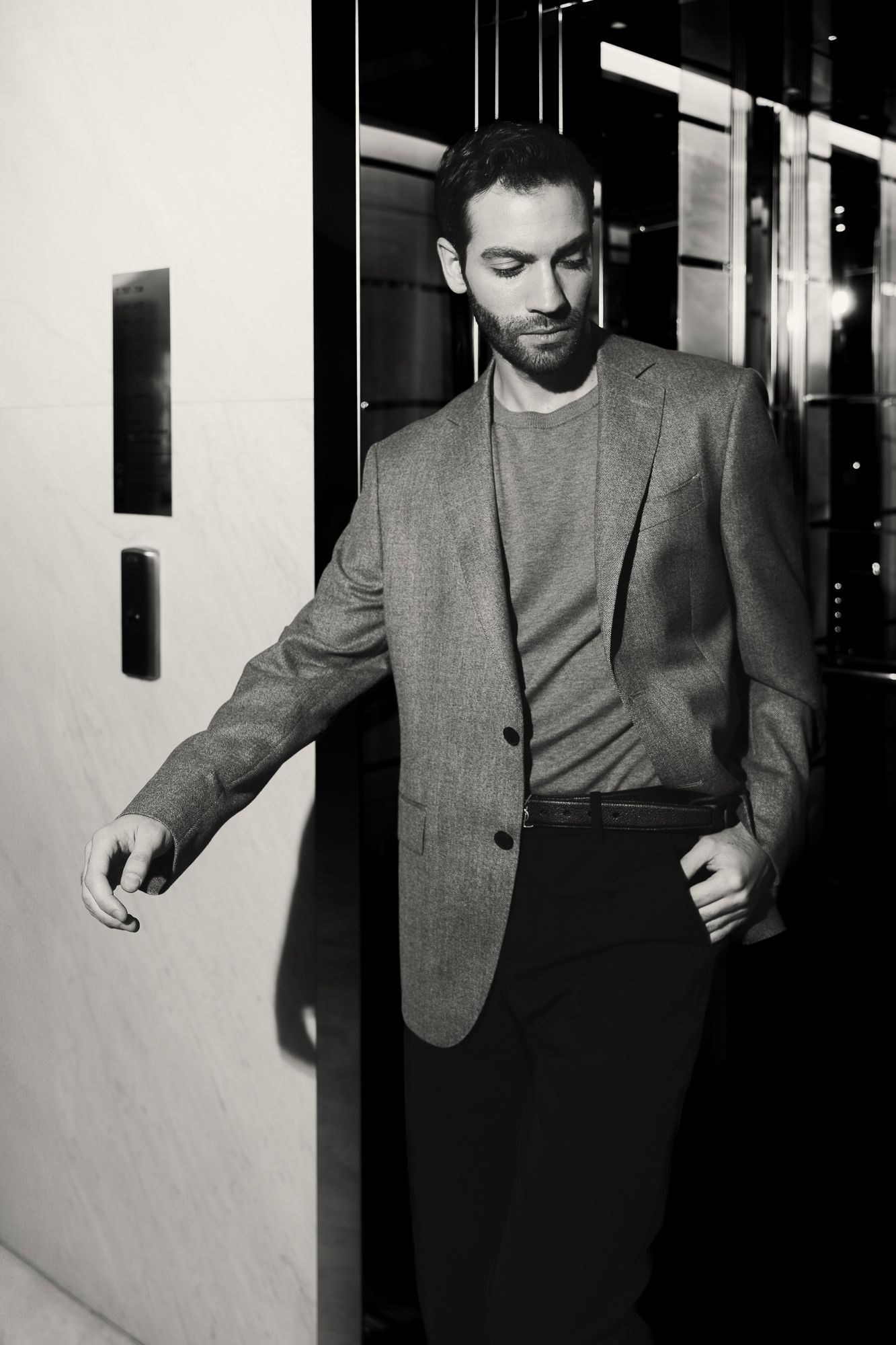 Stylish man in gray blazer posing near elevator doors in black and white photo.