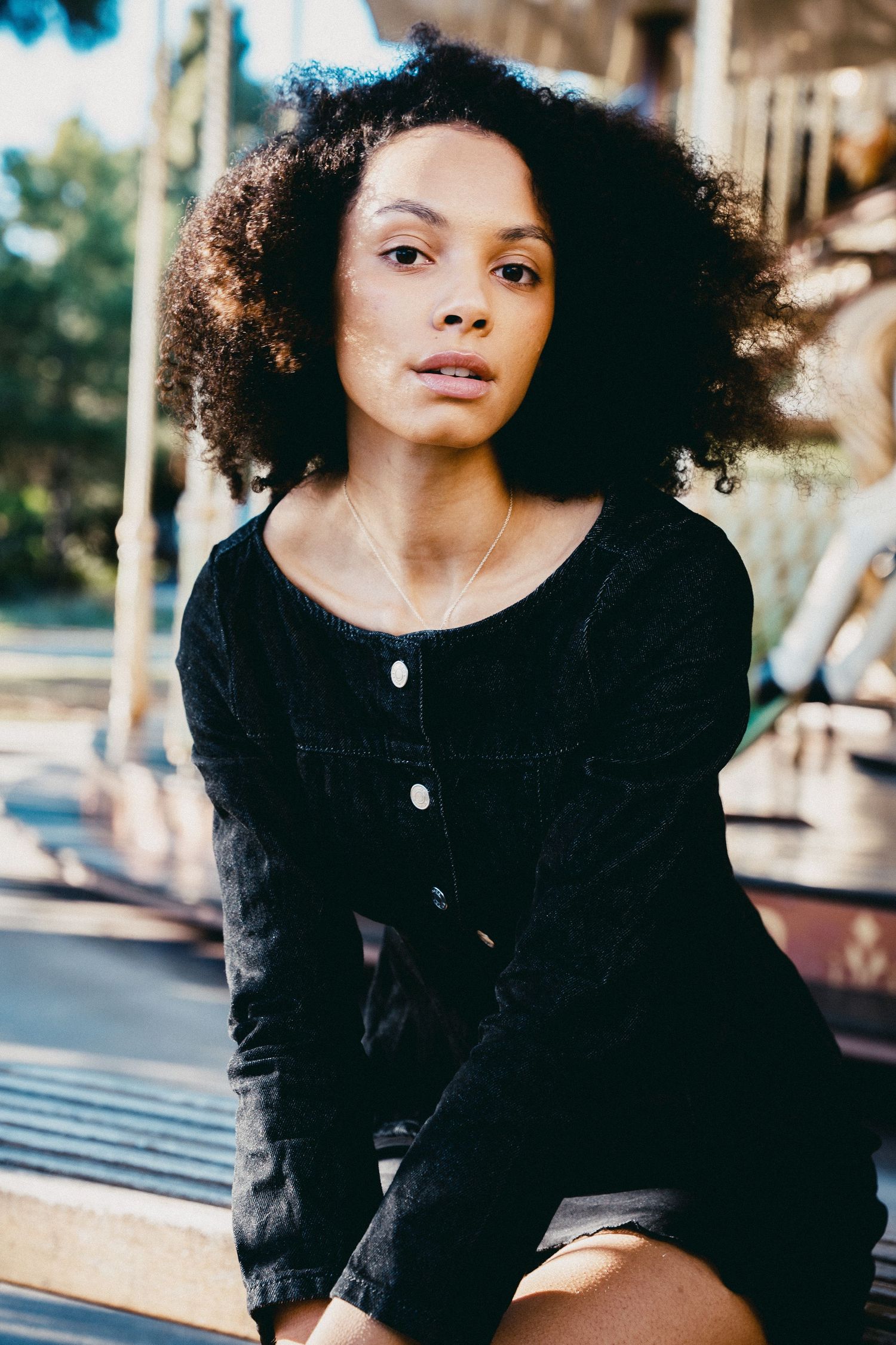 Young woman with curly afro hair in black denim dress sitting outdoors in sunlight.