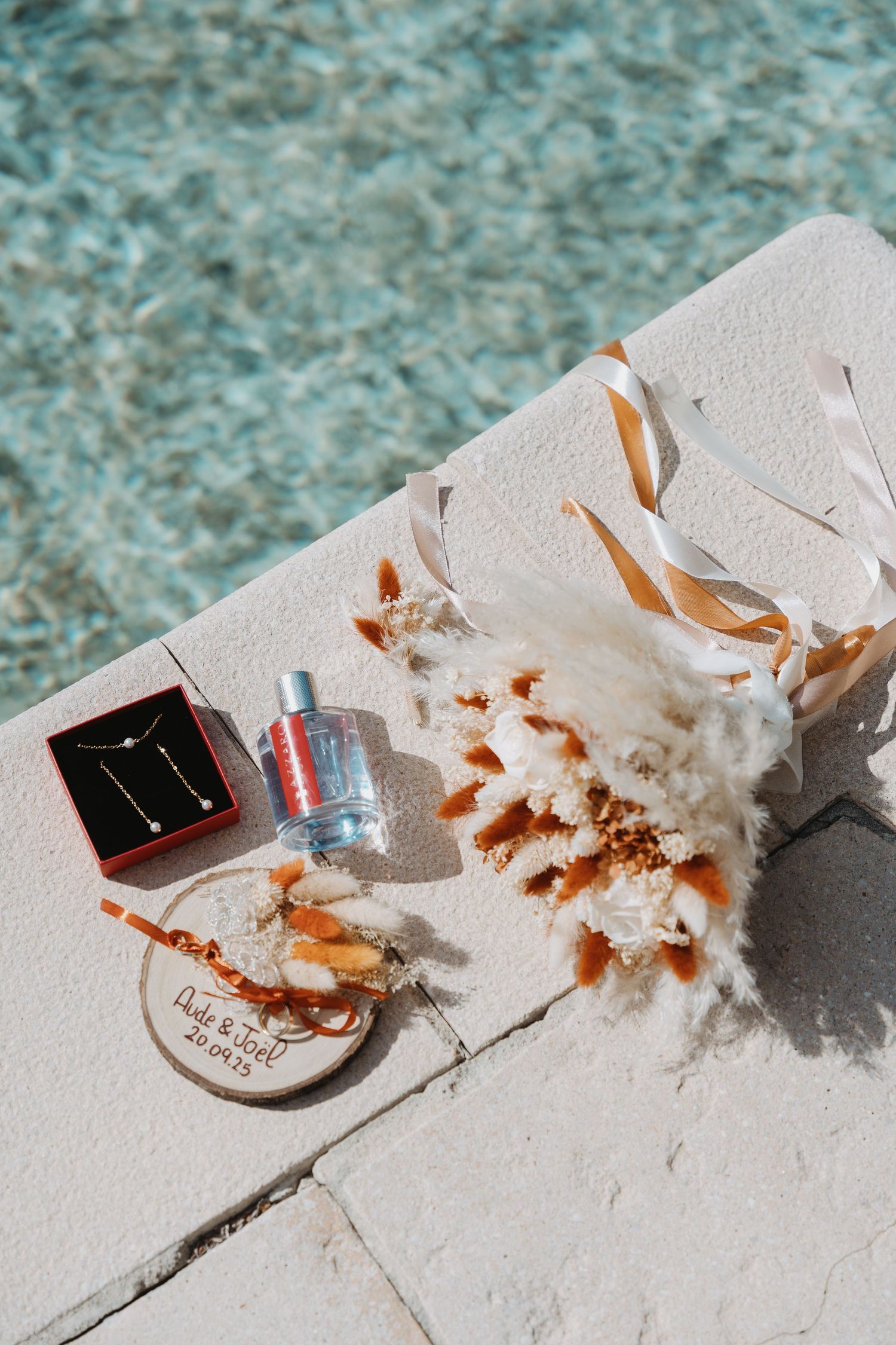Wedding flat lay with jewelry, perfume, and dried floral bouquet beside a turquoise pool.