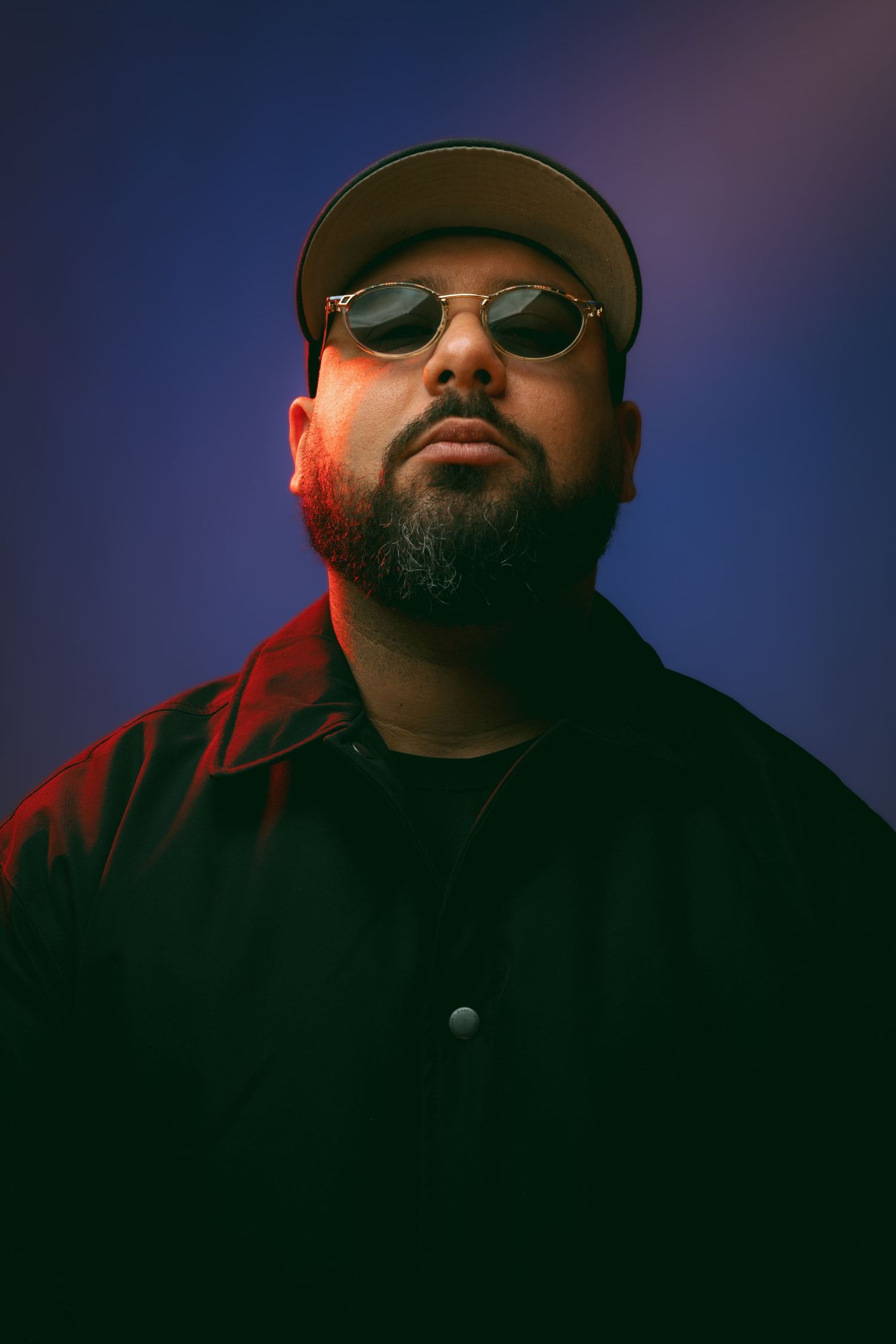 Heavyset bearded man wearing oval sunglasses and cap posing against blue-purple studio background.
