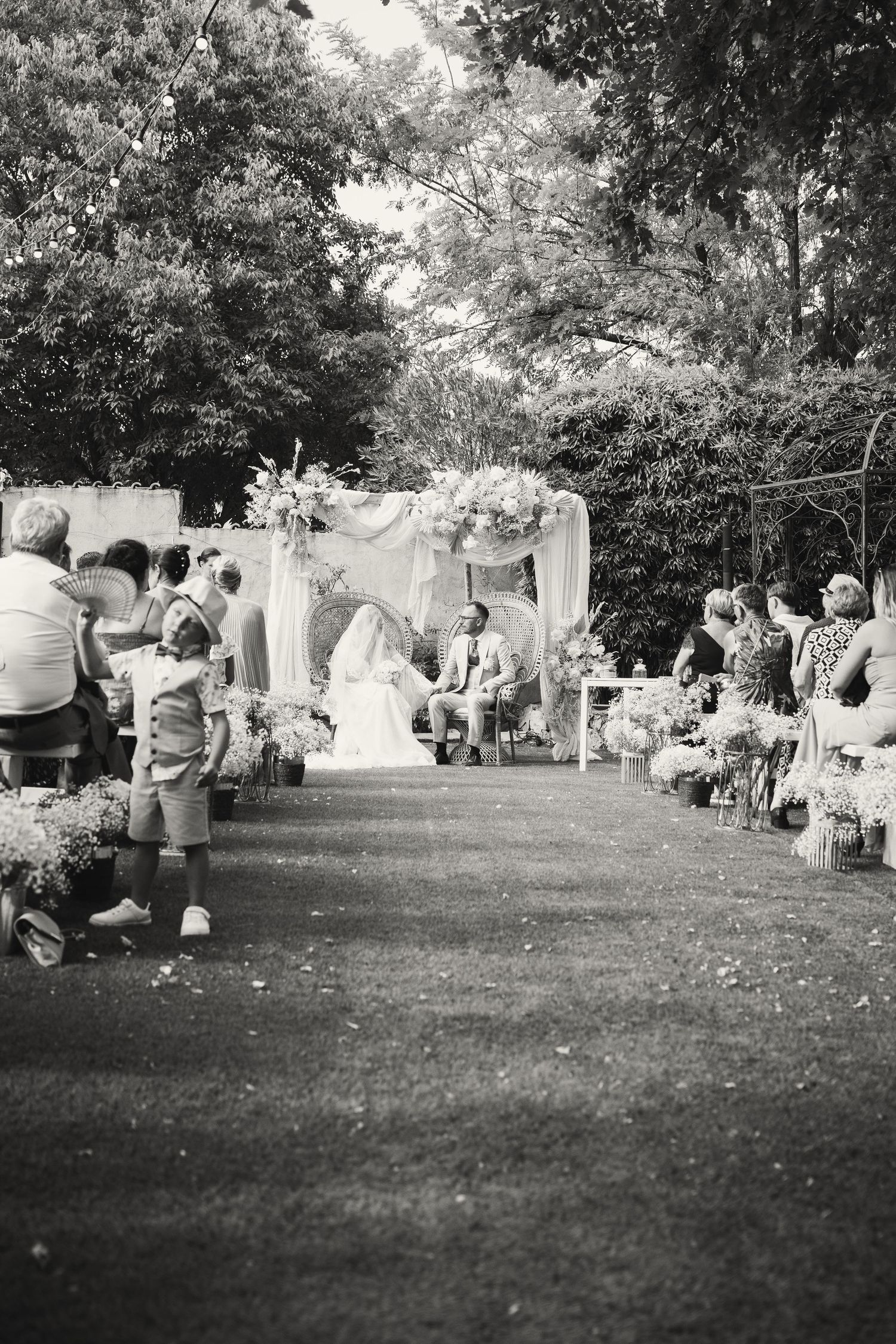 Black and white image of outdoor wedding ceremony with couple at floral altar and seated guests.