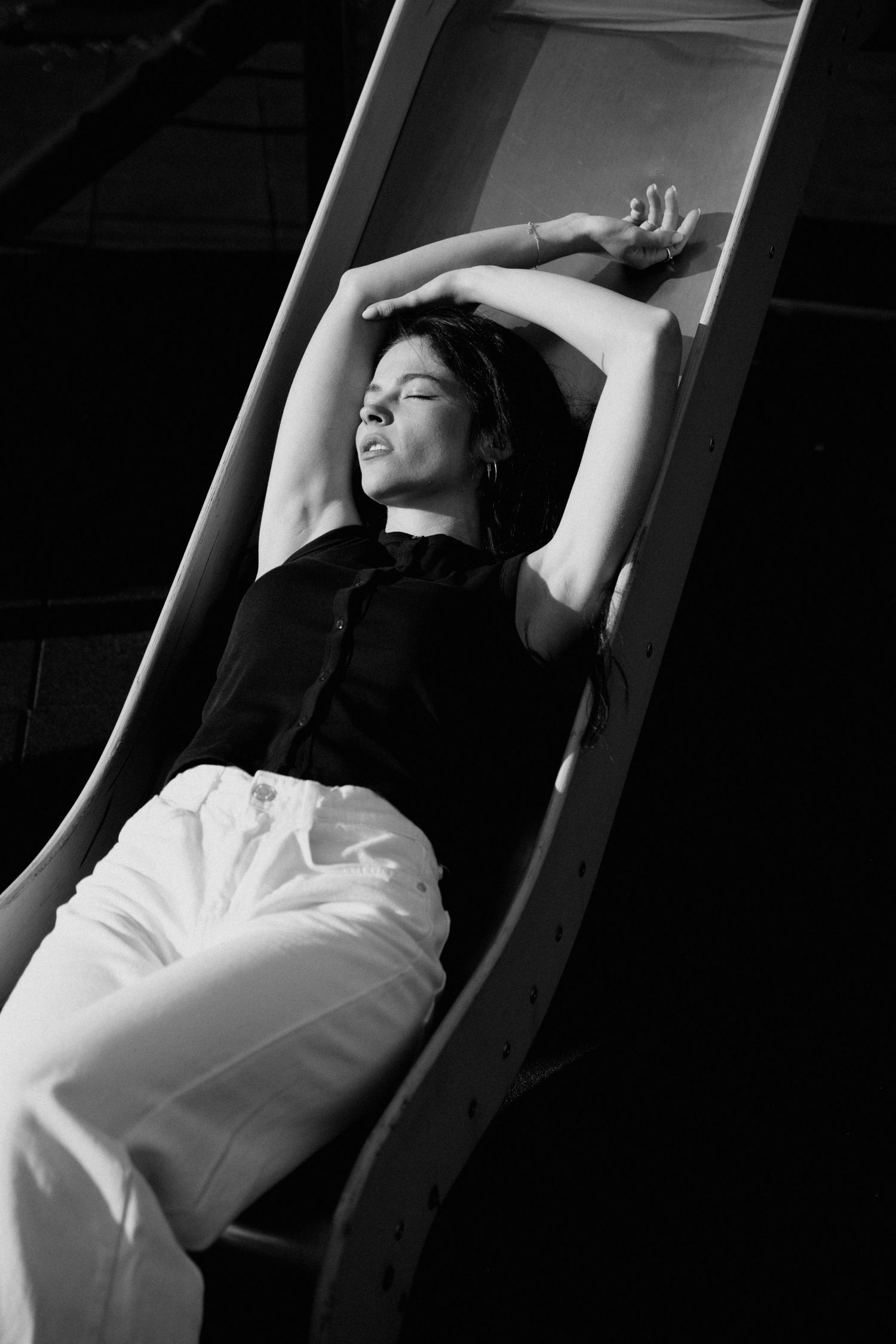 Black and white artistic photo of woman in black top and white pants reclining on wooden slide.