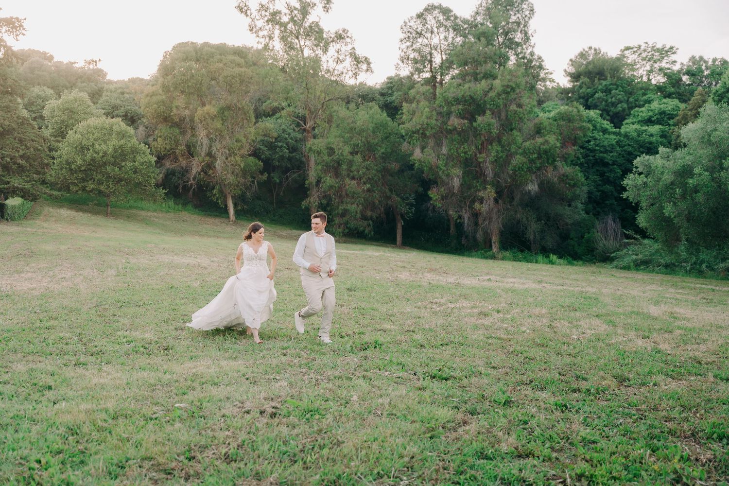 Bride and groom holding hands walking across green meadow surrounded by lush trees.