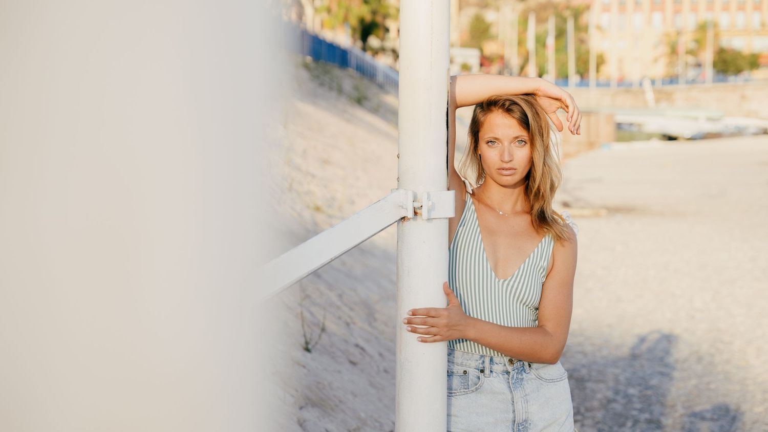 Young blonde woman in striped tank top leaning against white post on sunny beach promenade.