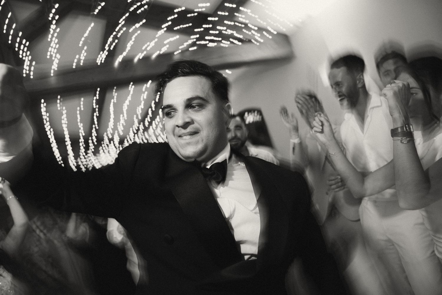 Groom in black tuxedo celebrating joyfully on the dance floor in a black and white wedding photo.