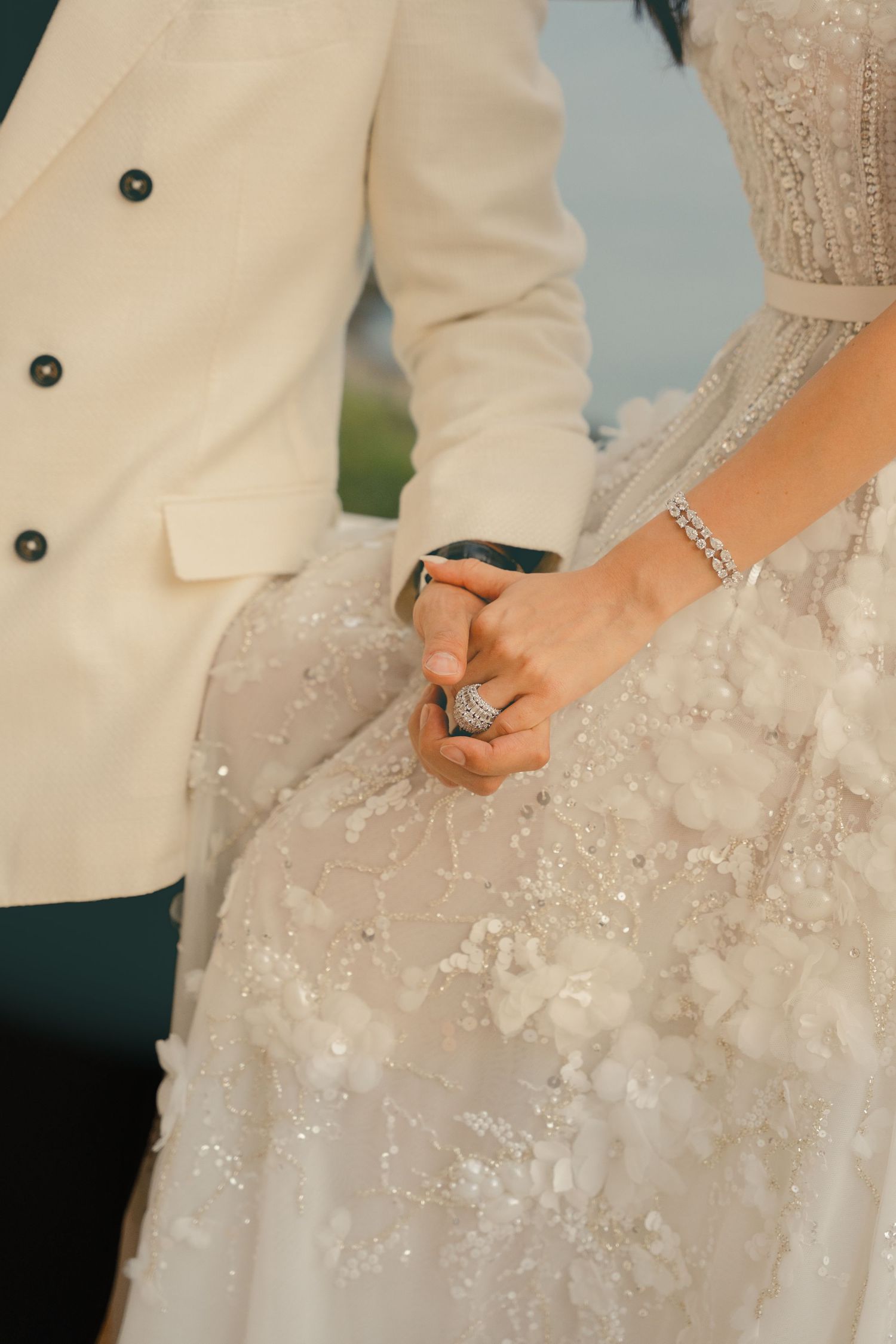 Bride and groom holding hands, showcasing a diamond ring and bracelet, wearing elegant white wedding attire.