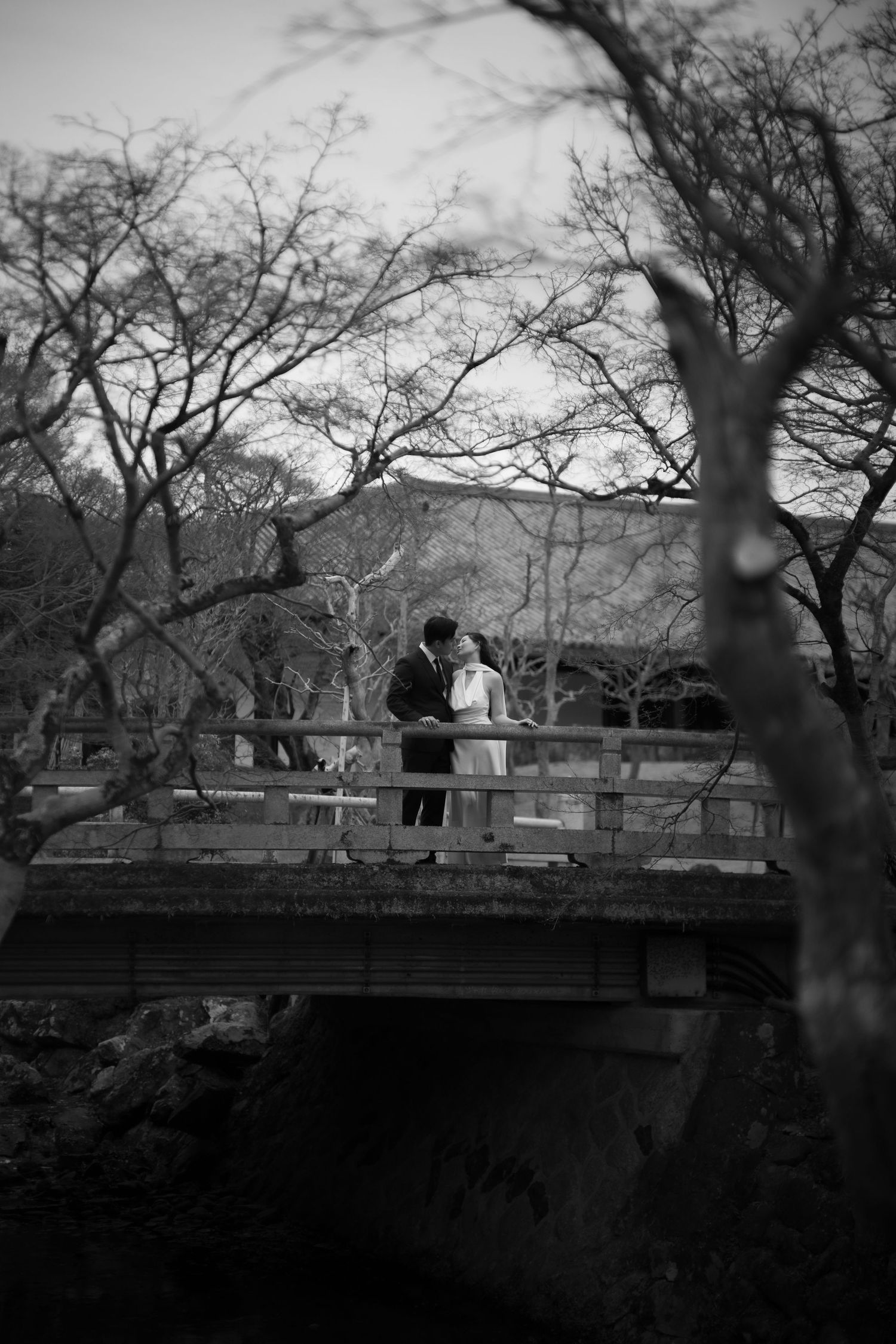 Black and white photo of a couple sharing a romantic kiss on a wooden bridge surrounded by bare trees.