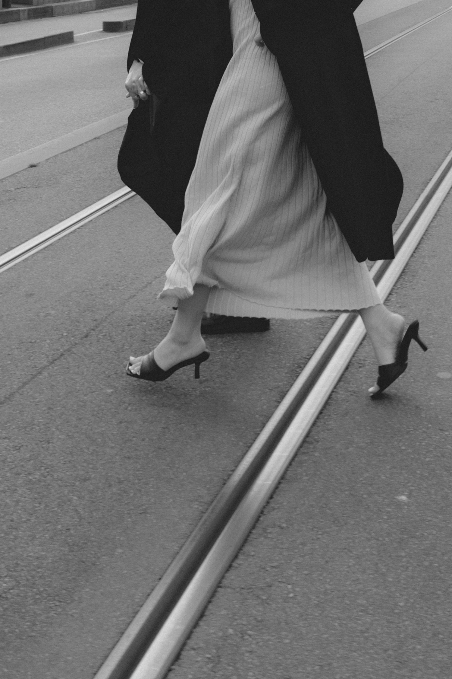 Black and white photo of a woman in a long pleated skirt and heeled mules crossing a street with tram tracks.