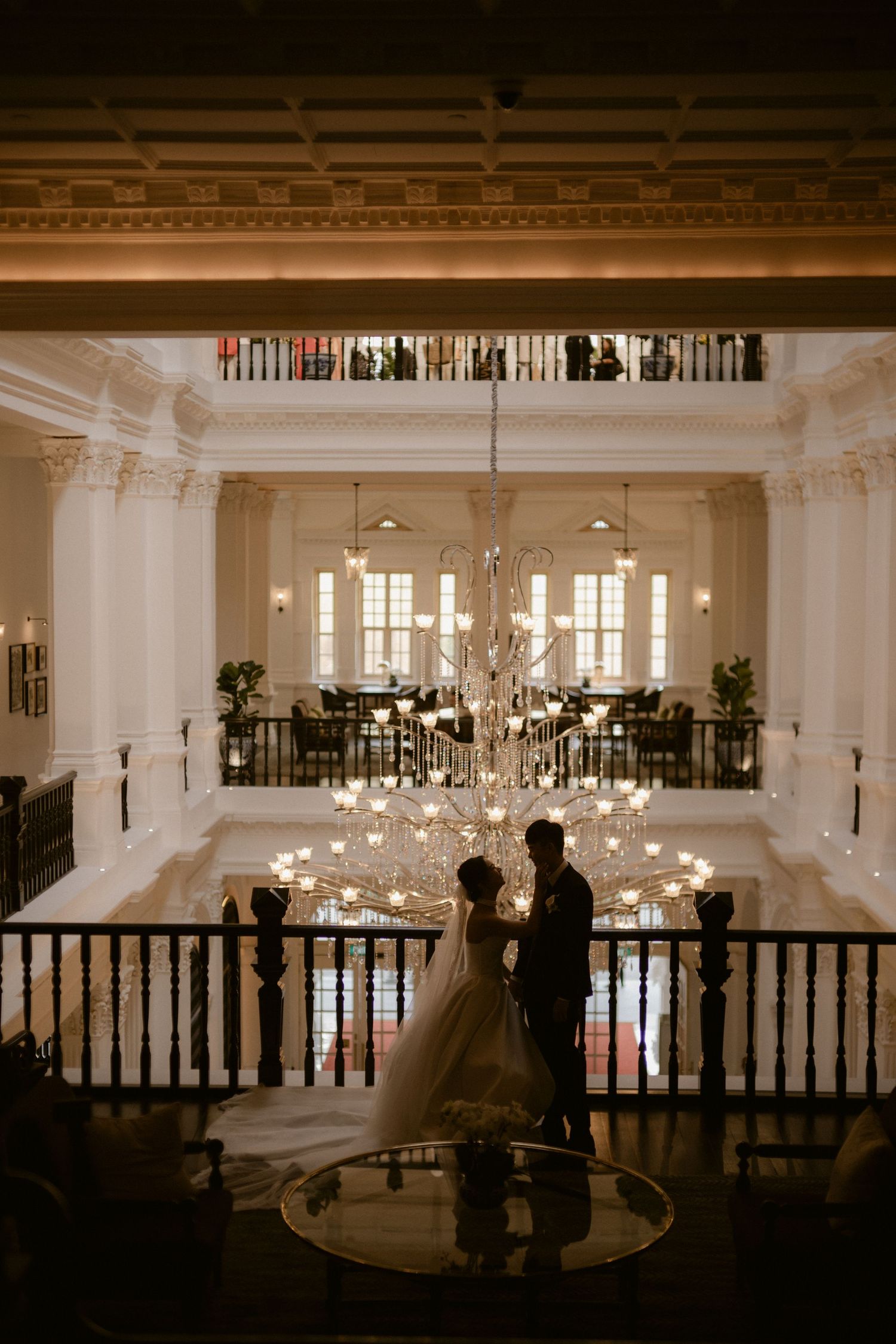 Bride and groom share romantic moment on grand hotel balcony with elegant chandelier and ornate white interior.