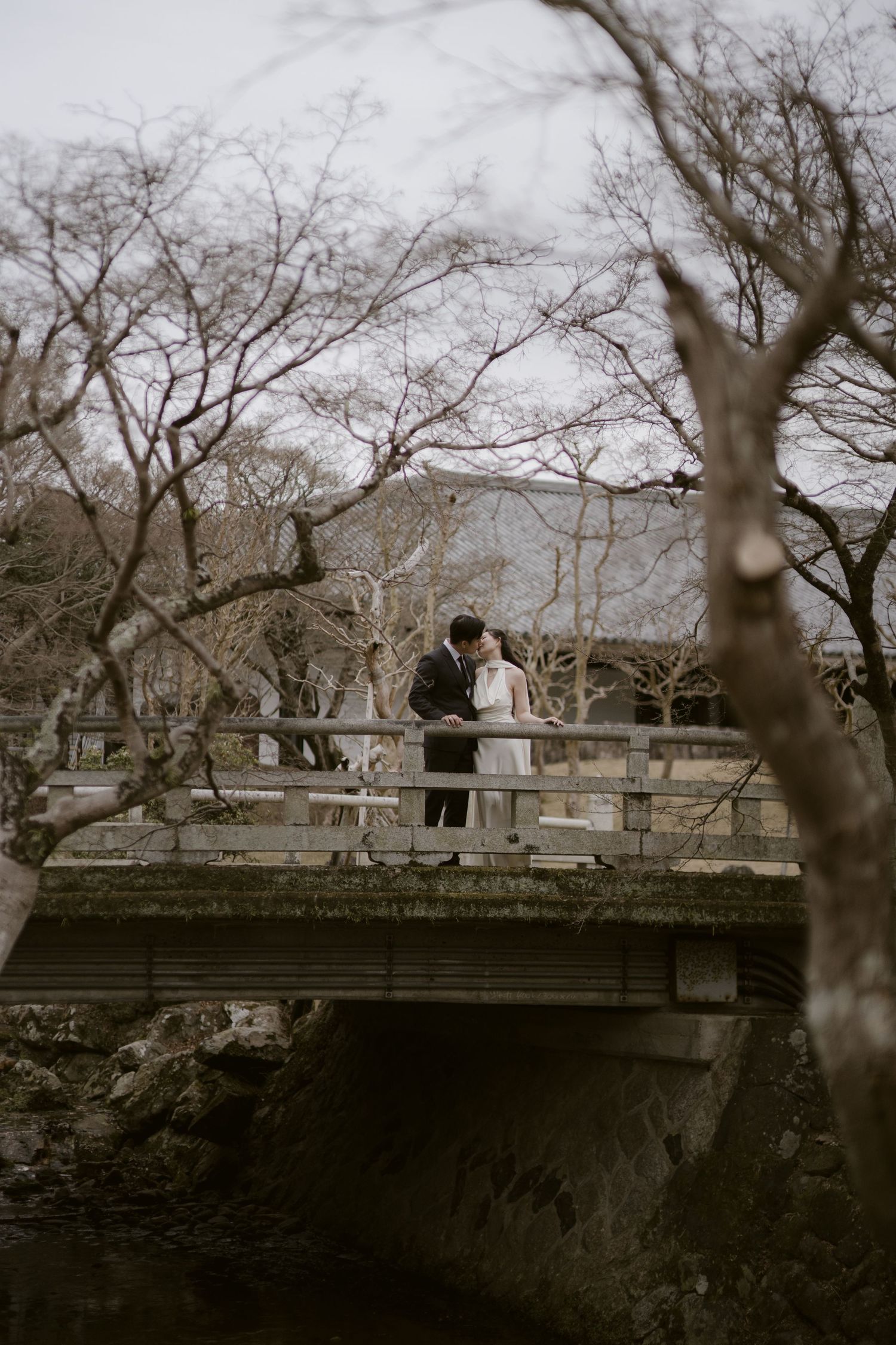 Couple sharing a romantic kiss on a moss-covered stone bridge surrounded by bare winter trees in a Japanese garden.
