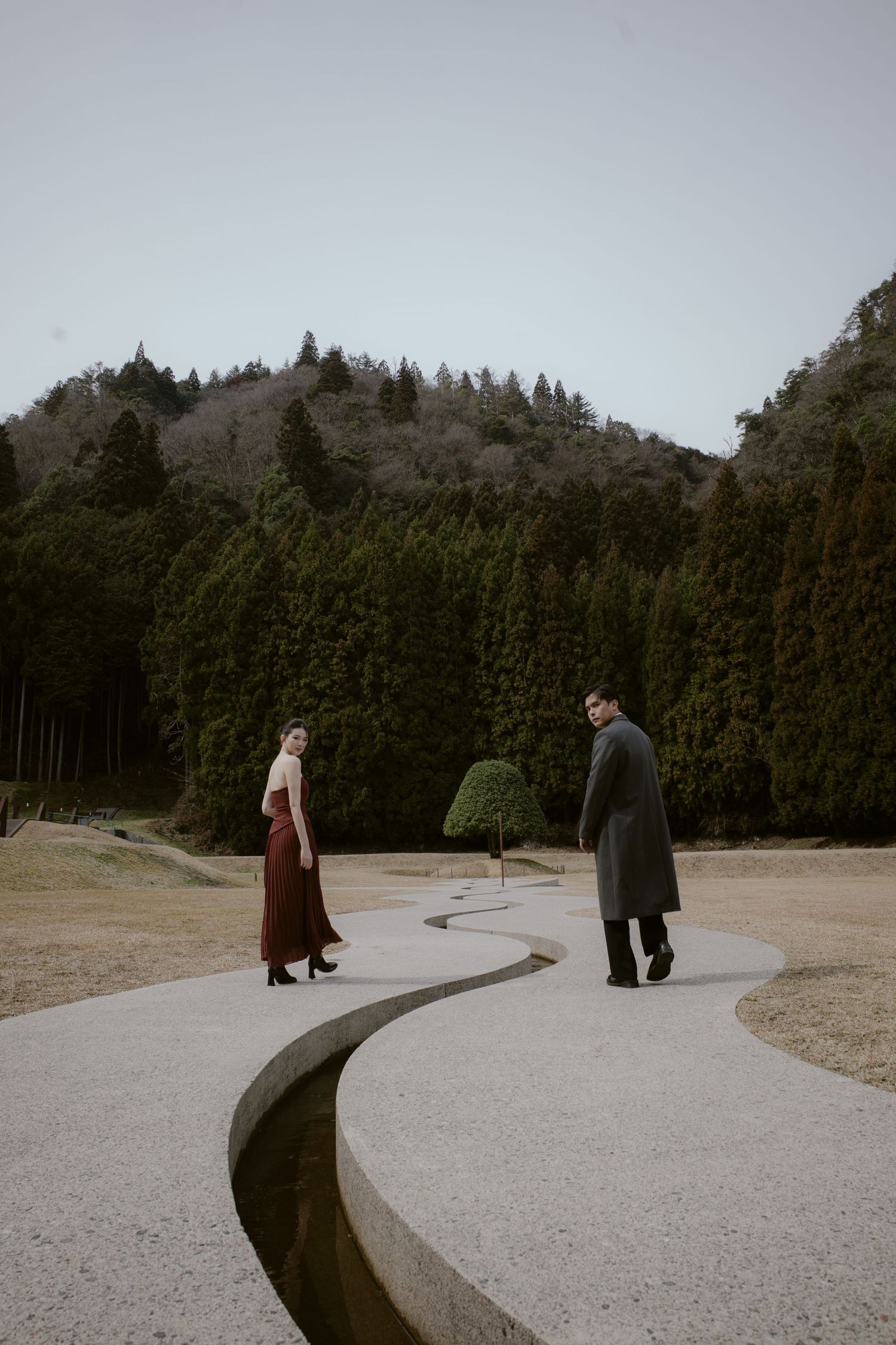 A couple in elegant attire walks along a curved stone path surrounded by lush green trees and a hillside.
