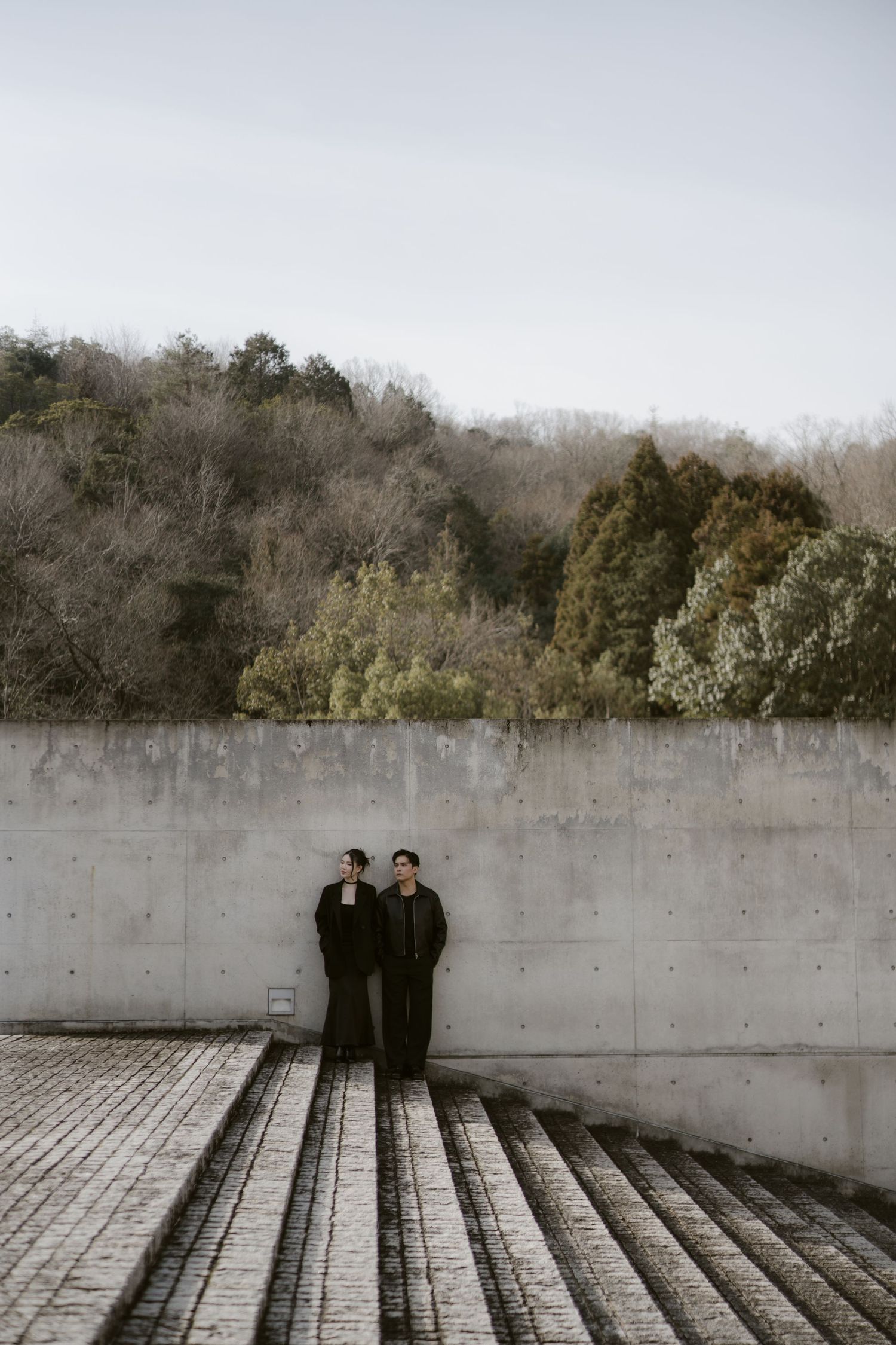 Two people in black outfits stand against a concrete wall with lush trees and overcast sky in the background.