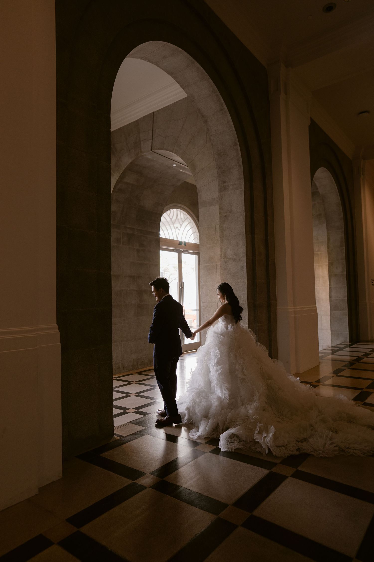 Bride in flowing white gown holds groom's hand walking through elegant arched hallway with checkered marble floors.