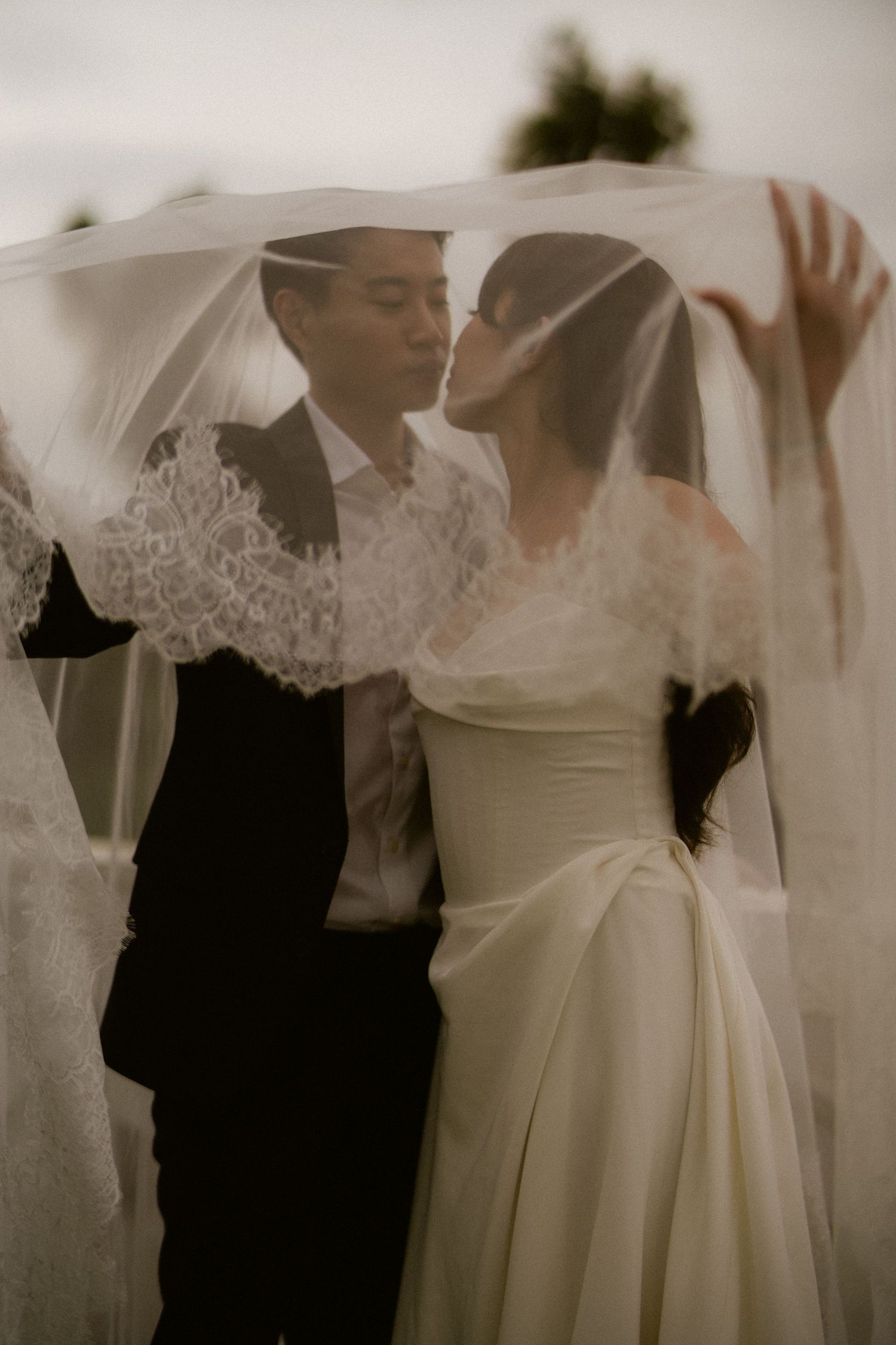 A bride and groom share a romantic kiss under a flowing lace wedding veil outdoors.