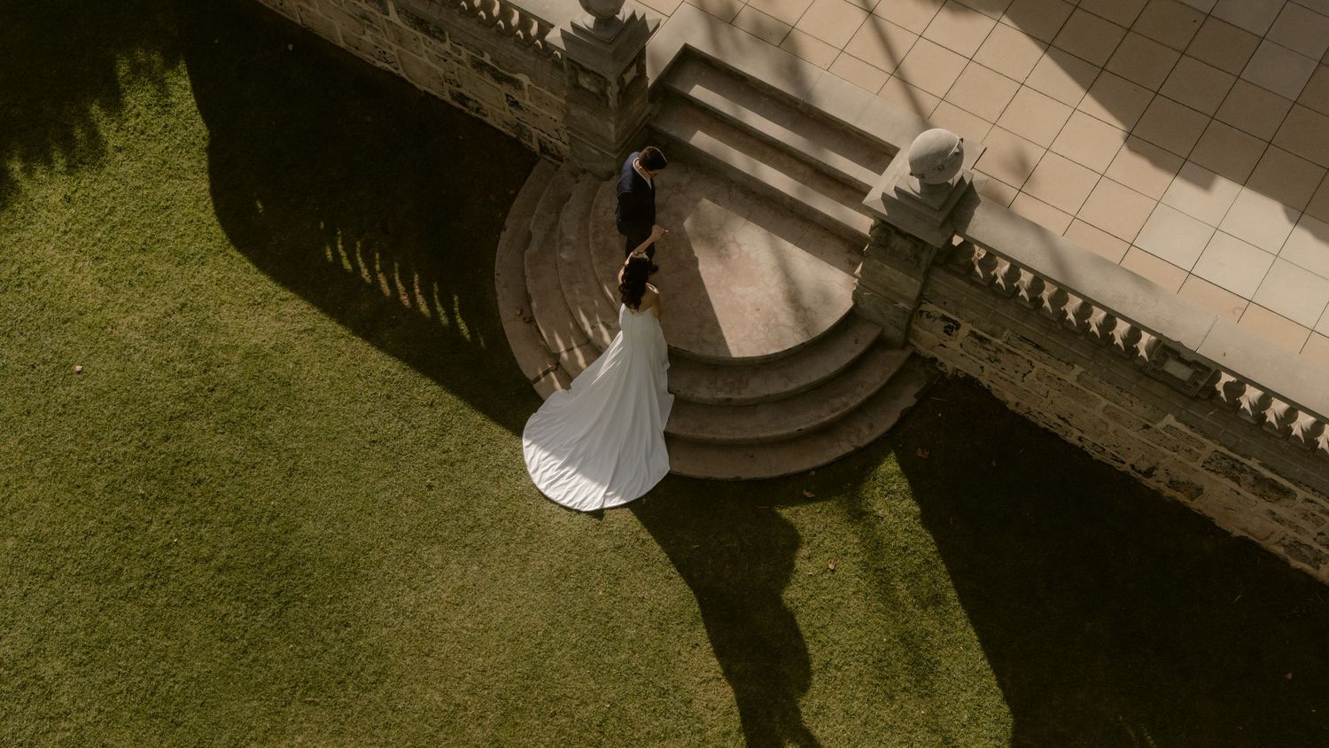 Aerial drone view of a bride and groom walking up stone steps at a wedding venue surrounded by lush green lawns.