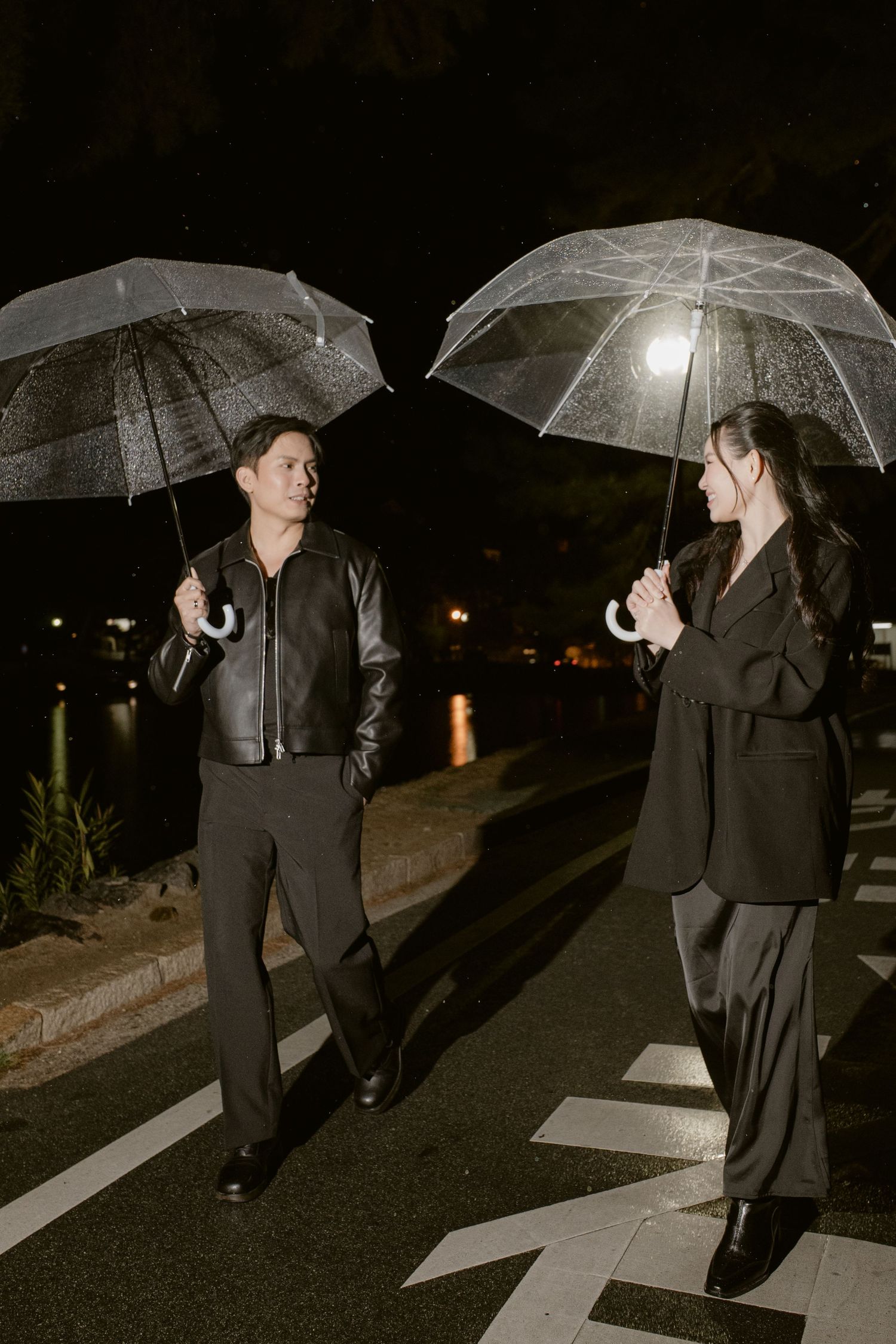 A man and woman holding clear umbrellas stand on a crosswalk at night, exchanging glances in the rain.