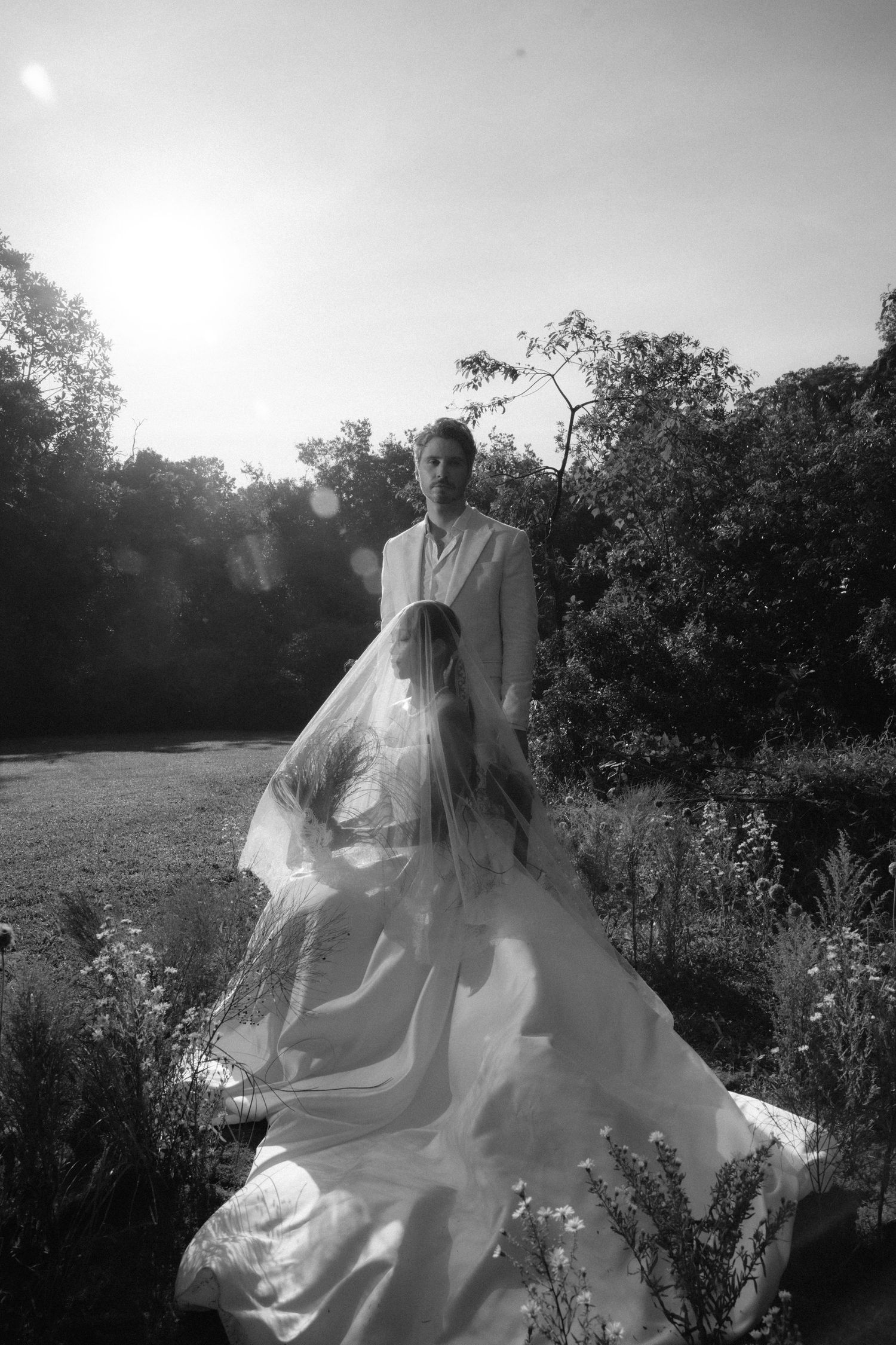 Black and white photo of a bride in flowing gown and veil with groom standing in sunlit garden.