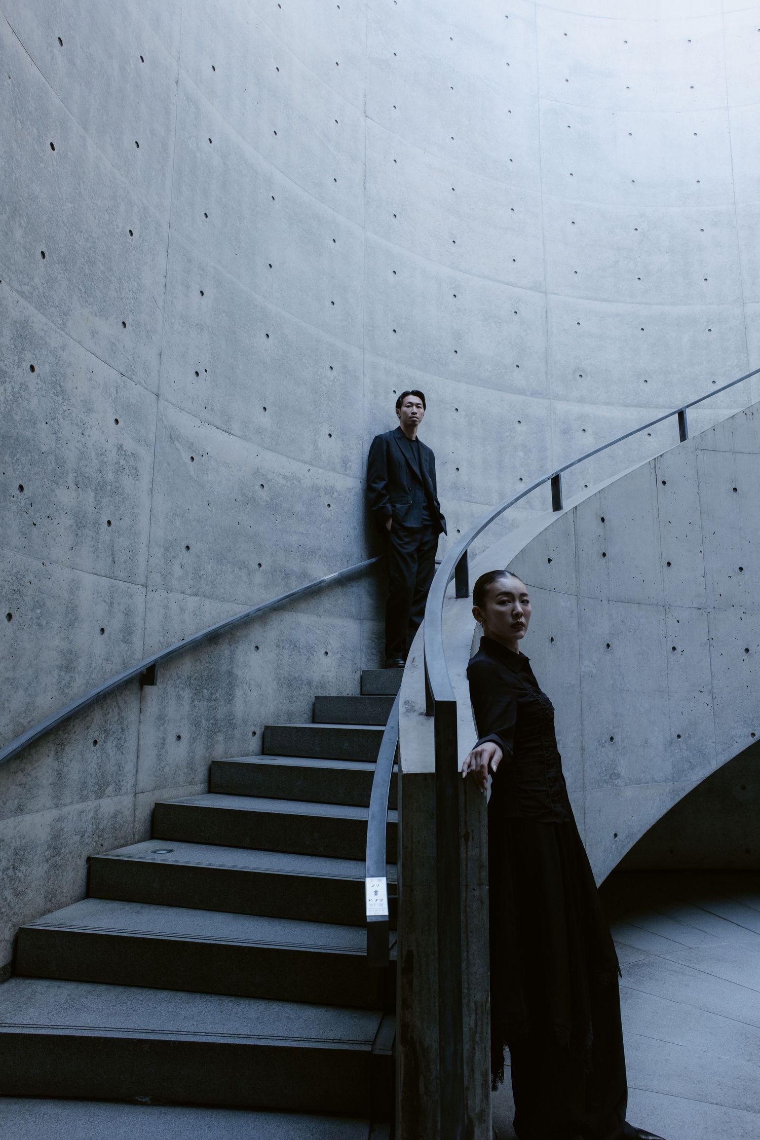 Two elegantly dressed figures pose on a curved concrete staircase in a modern architectural interior.