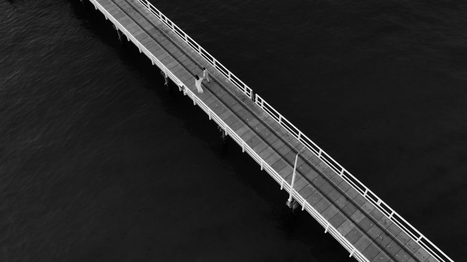 Aerial black and white photo of a long narrow bridge stretching over dark water.