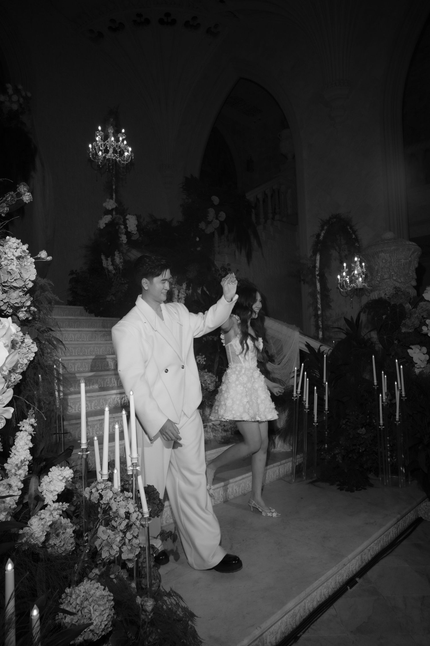 Black and white photo of a couple in formal attire sharing a moment at an elegant candlelit venue with floral arrangements.