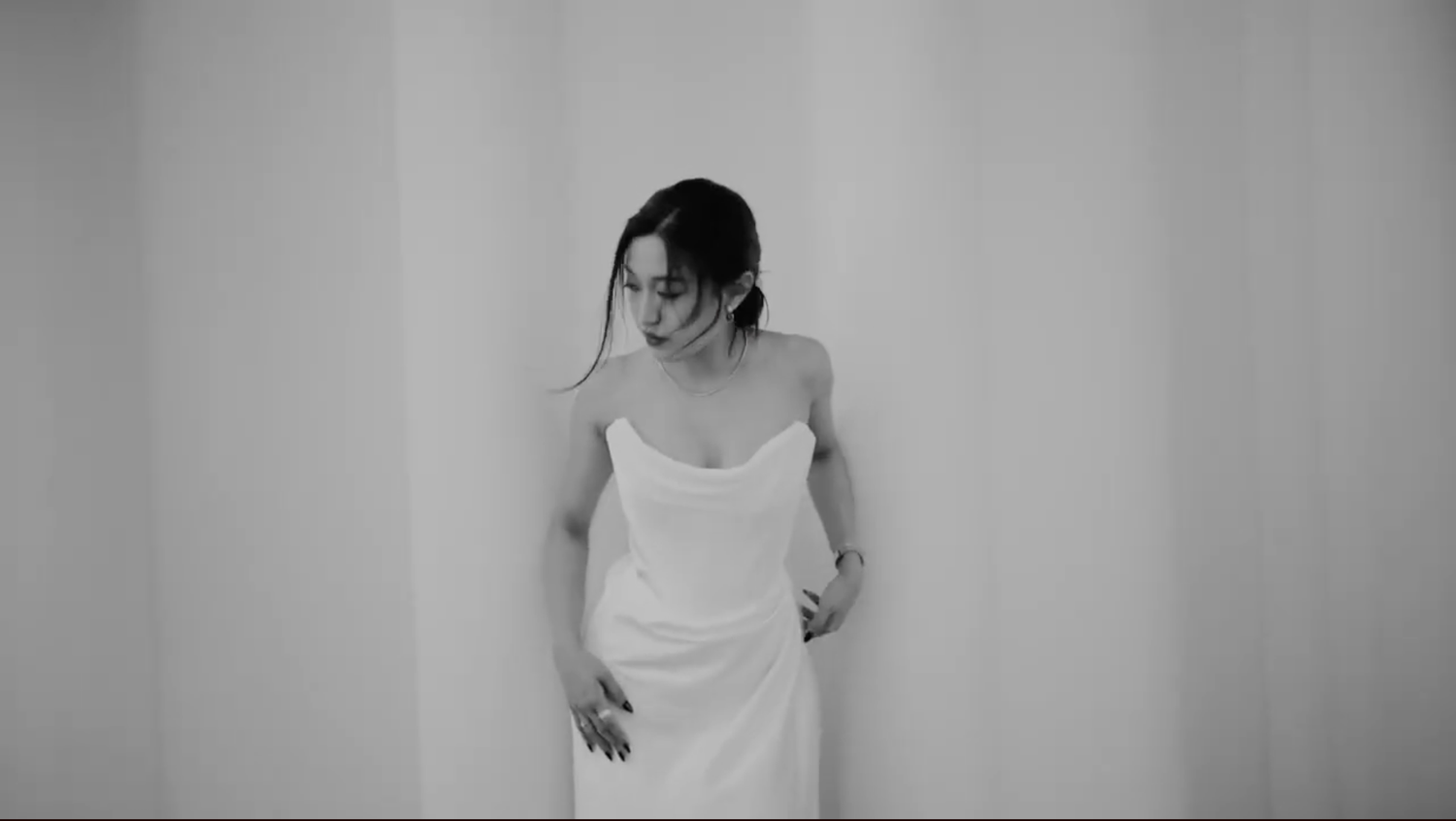 Black and white photo of a woman in a white slip dress posing gracefully against a light wall.