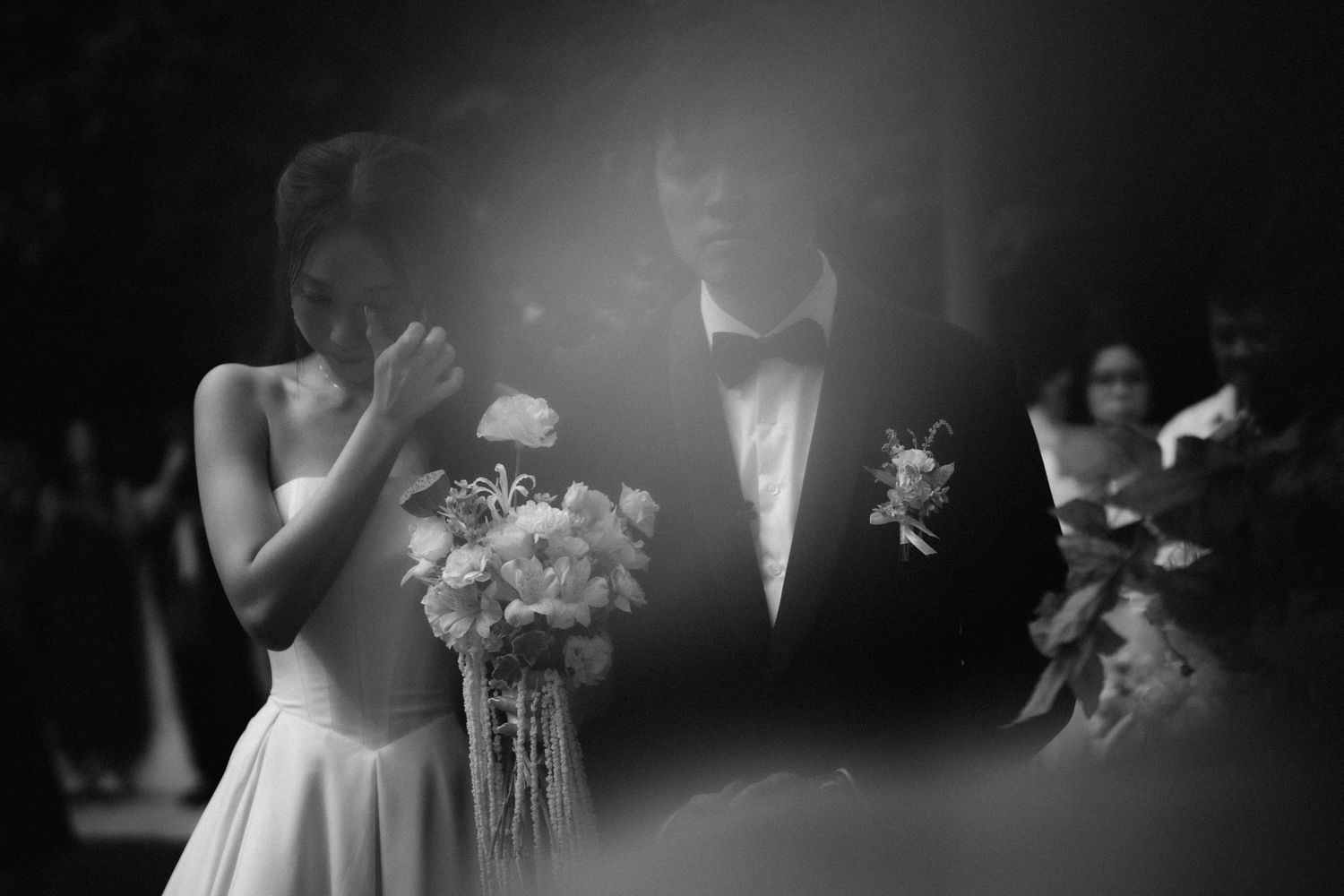 Black and white wedding photo of bride holding bouquet and groom in tuxedo during outdoor ceremony.