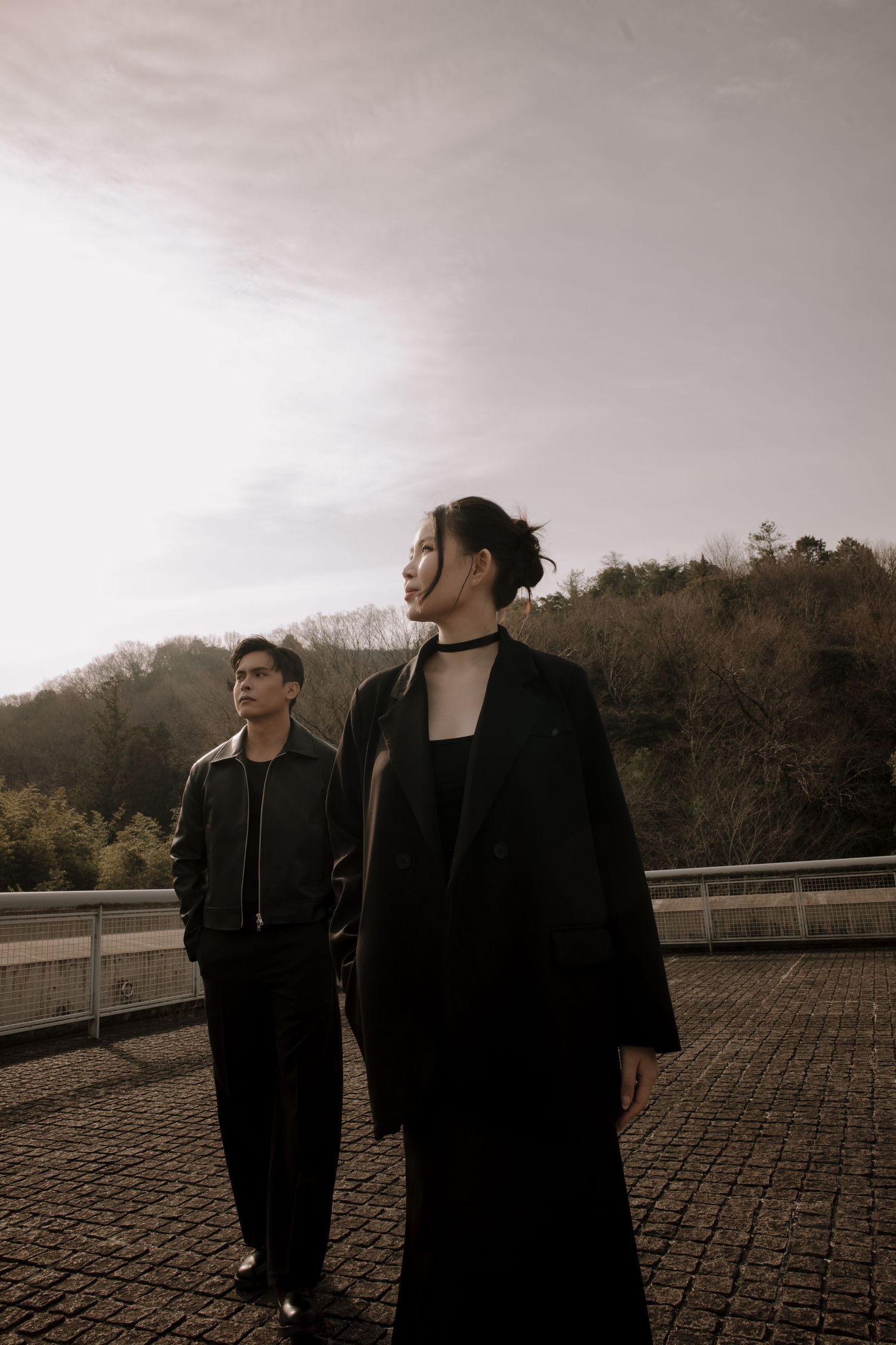 Two stylish individuals in dark outfits stand on a rooftop terrace with autumn trees and overcast sky.