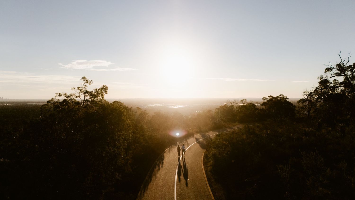 Two silhouetted figures walk down a sunlit road surrounded by lush trees at golden hour.