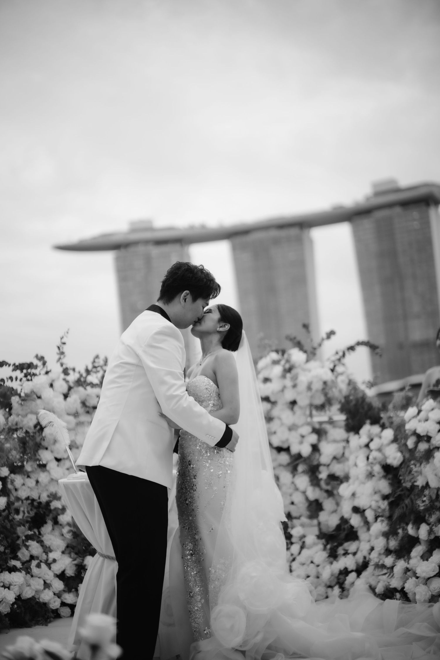 Black and white photo of a couple sharing a kiss at their wedding ceremony with Marina Bay Sands in the background.
