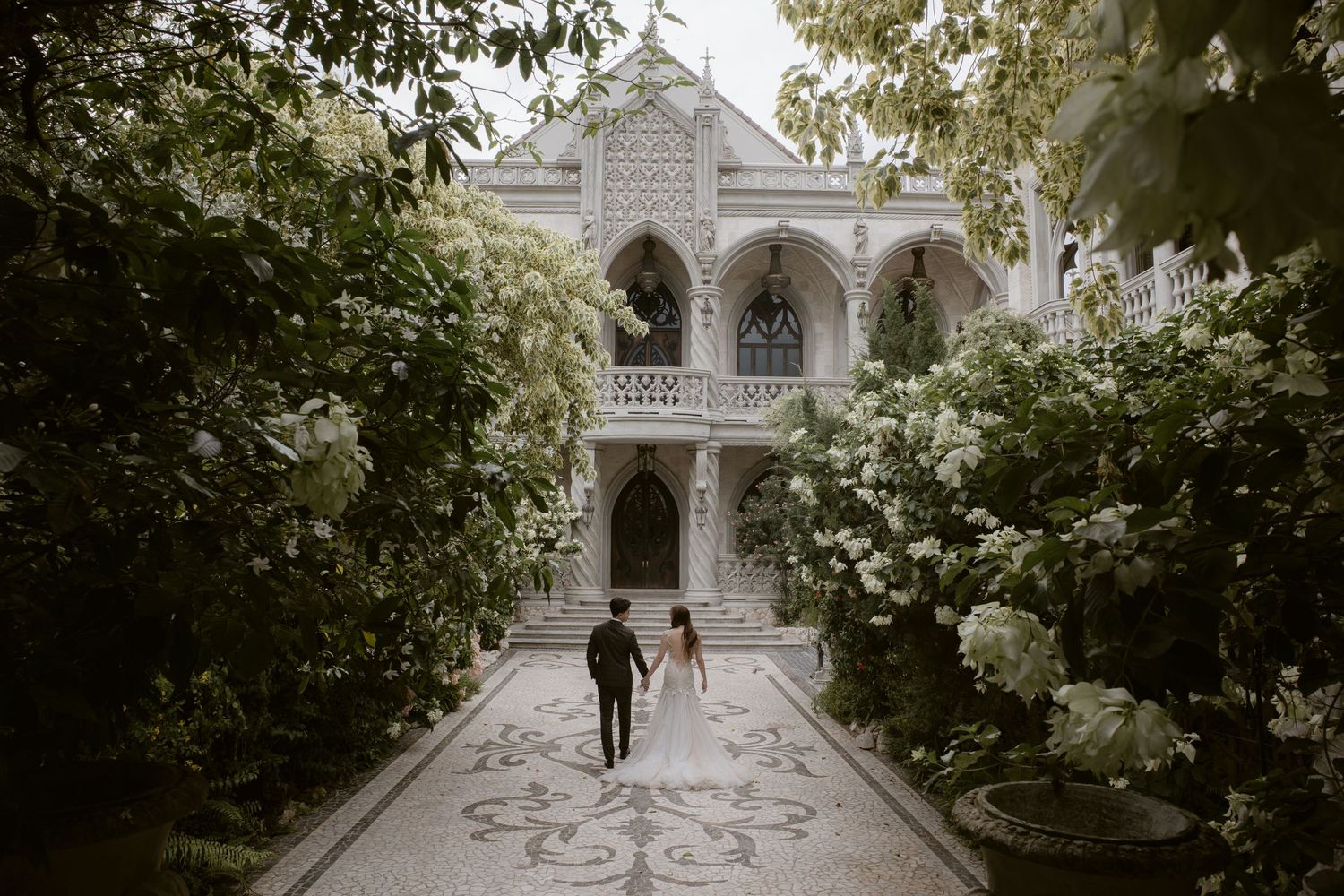 Couple walks hand-in-hand toward ornate white Gothic mansion surrounded by lush green trees and blooming white flowers.
