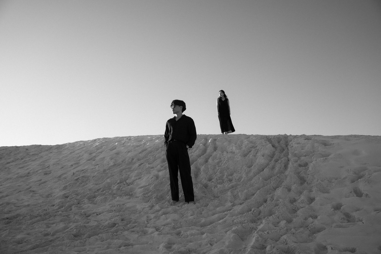 Two figures in dark clothing stand apart on a sandy dune under a clear sky in a black and white photo.