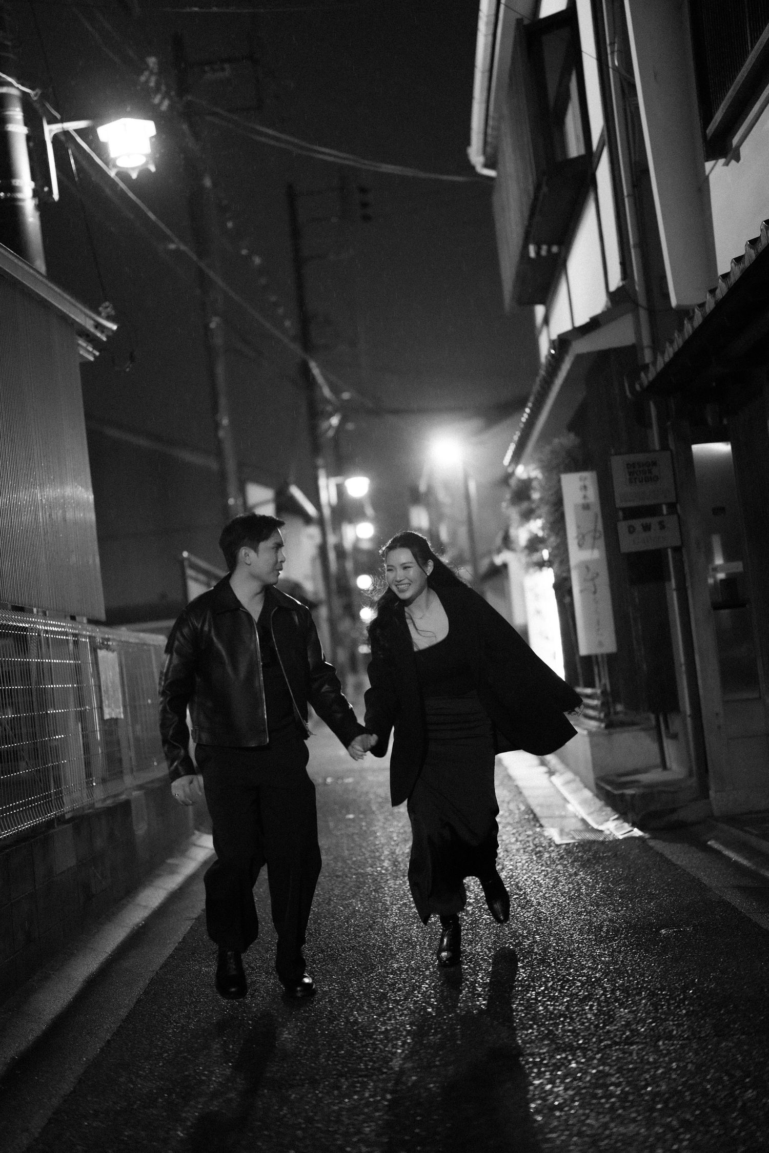 Black and white photo of a couple holding hands walking down a narrow Japanese alley at night.