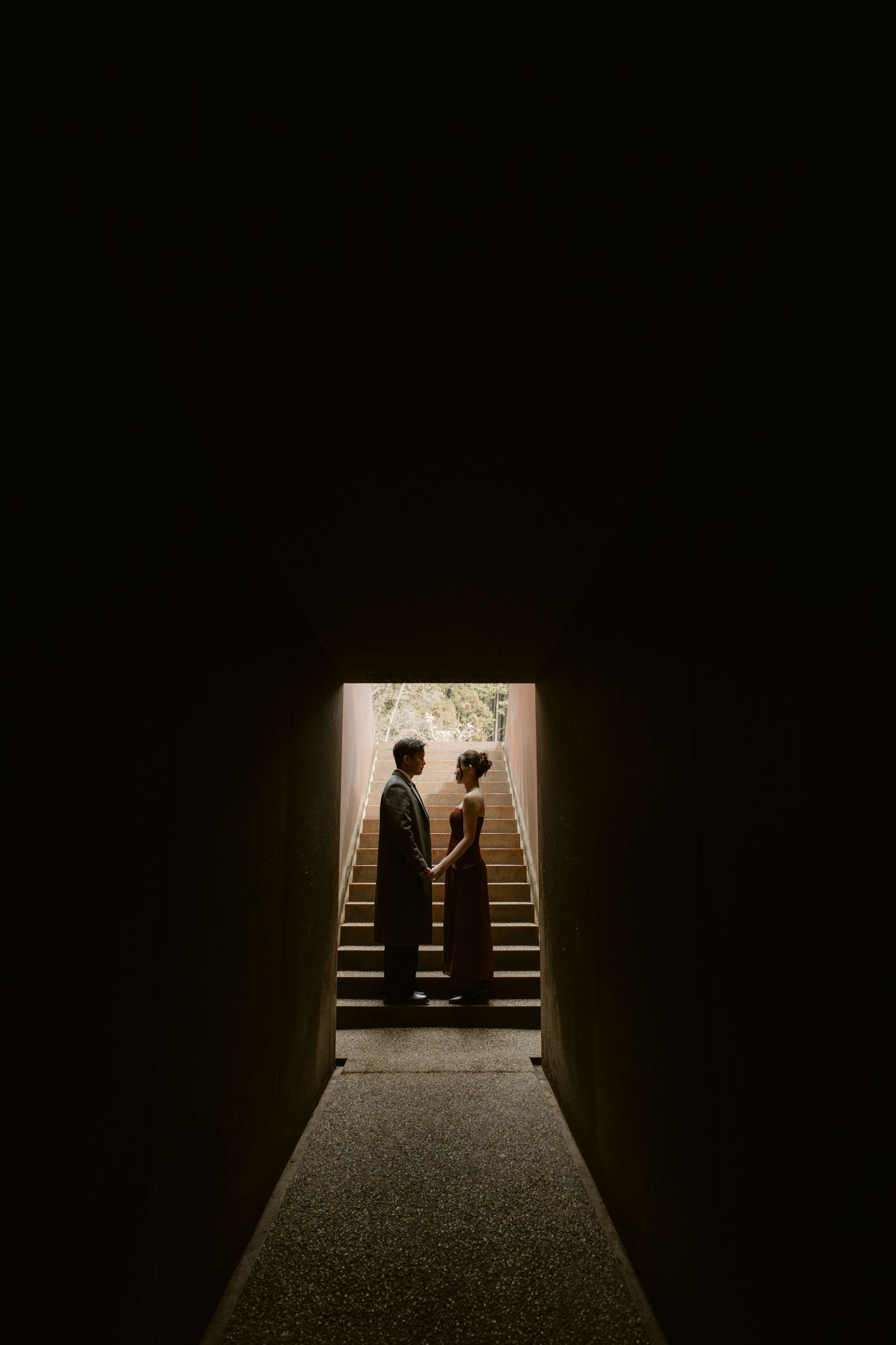 Couple holding hands in a dimly lit hallway framing a staircase, creating a romantic silhouette effect.