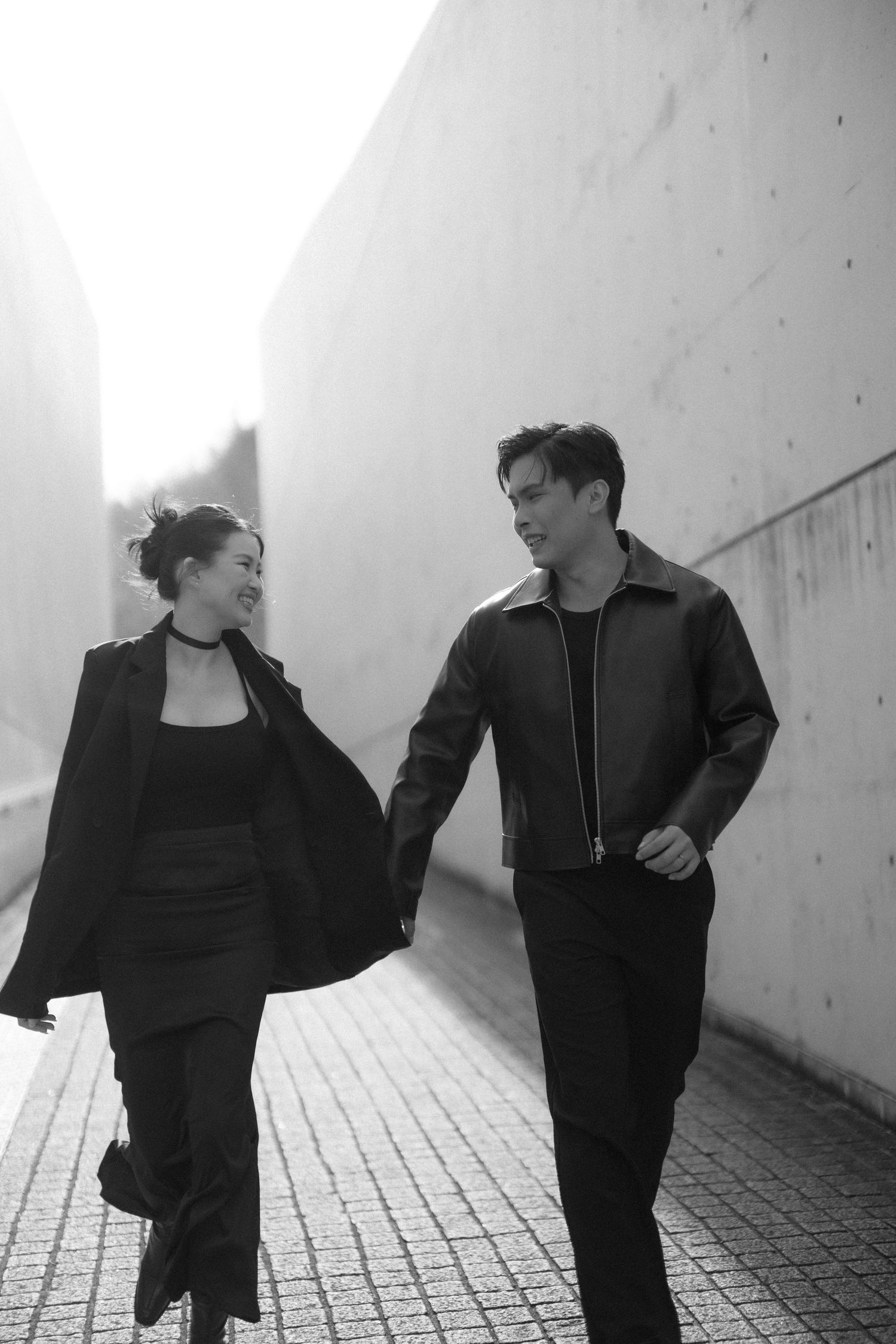Black and white photo of a stylish couple walking together, smiling at each other near a concrete wall.