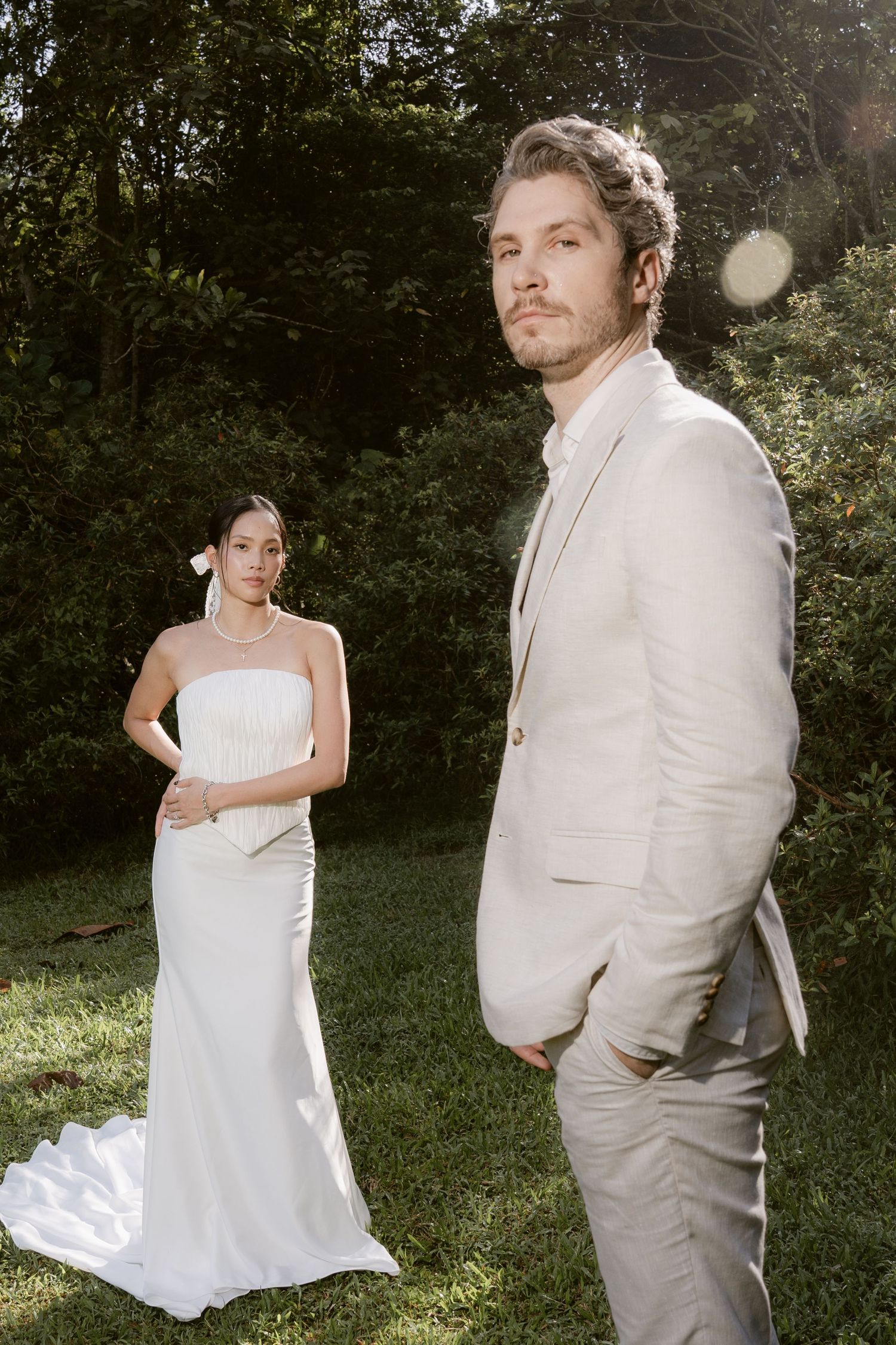Bride in white strapless gown and groom in beige suit pose outdoors on grass with lush green trees behind them.