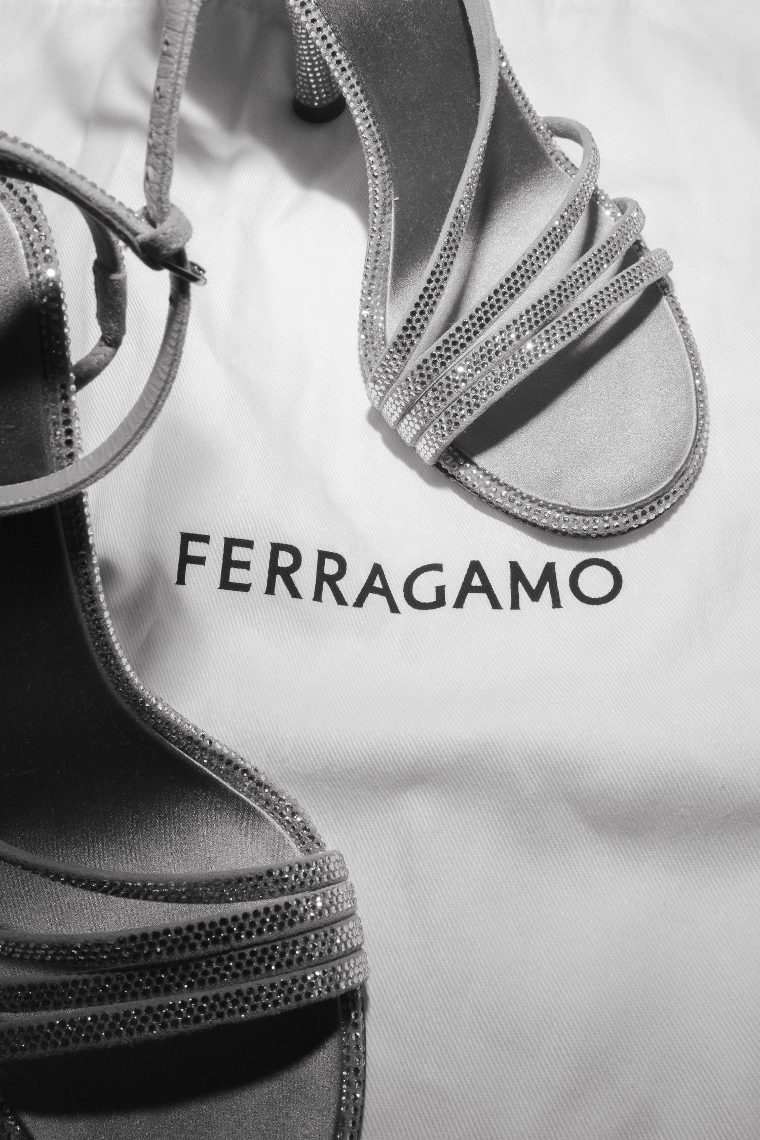 Ferragamo crystal-embellished strappy heeled sandals displayed on a white dust bag in black and white.