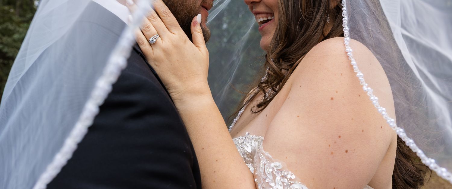 A bride and groom share a romantic moment under a wedding veil, with the bride smiling and showing her engagement ring.