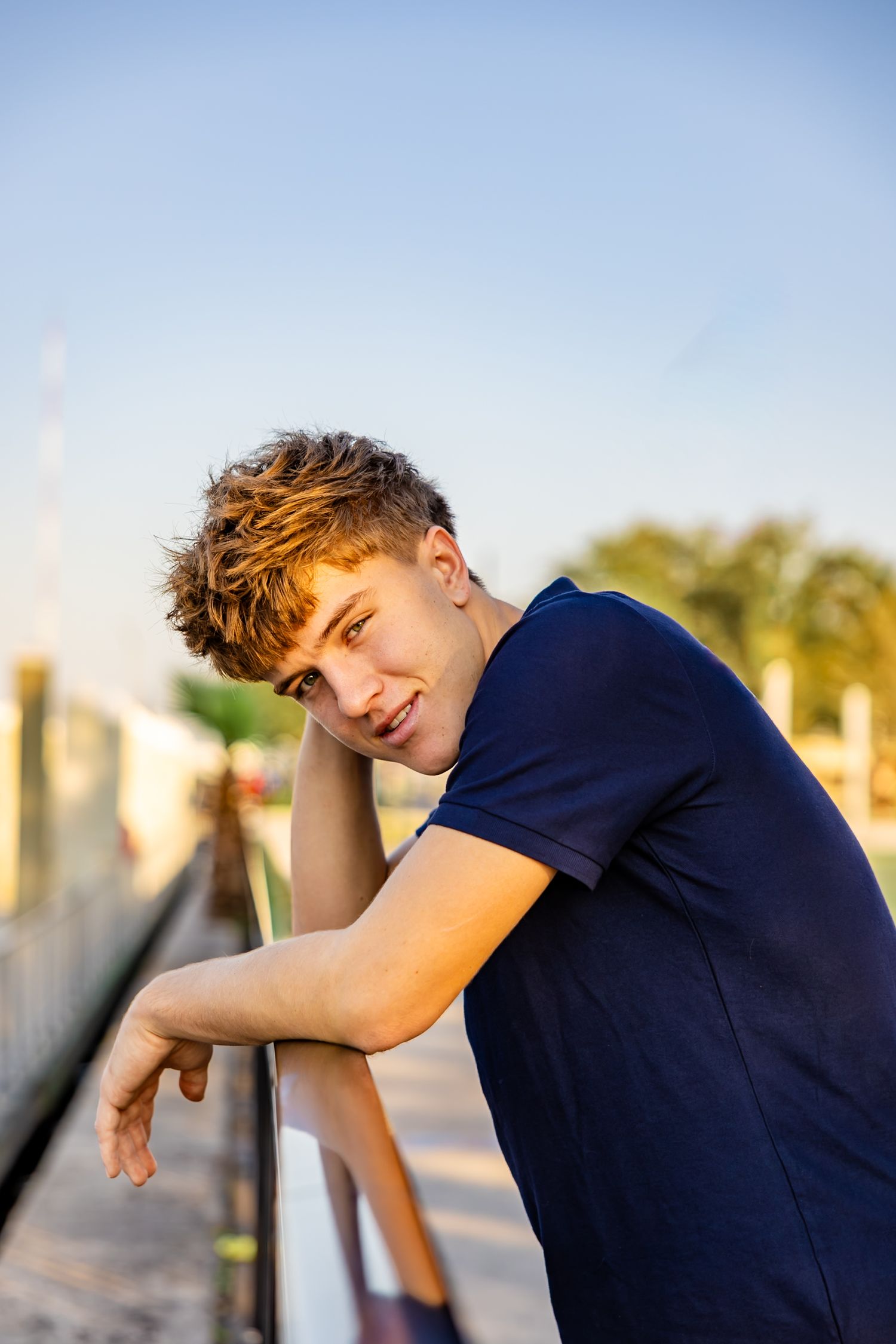 Senior guy leaning on a railing outdoors posing for senior photos during golden hour.