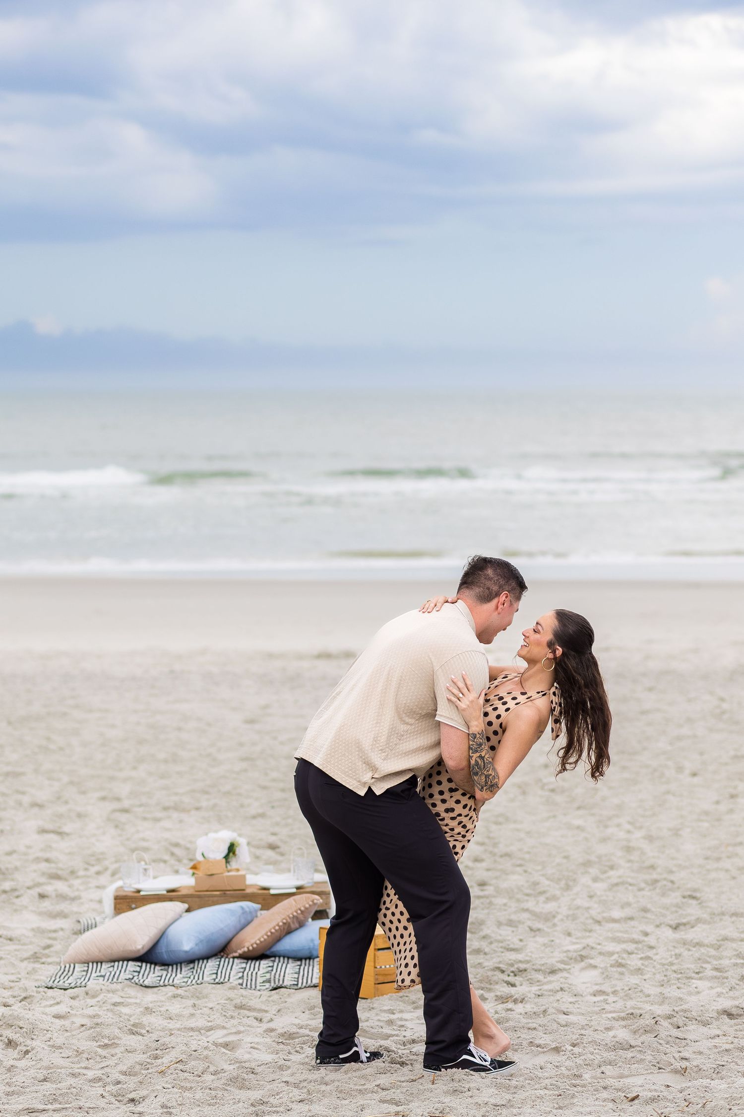 Couple sharing a romantic dip kiss after man proposes on a sandy beach with a picnic setup.