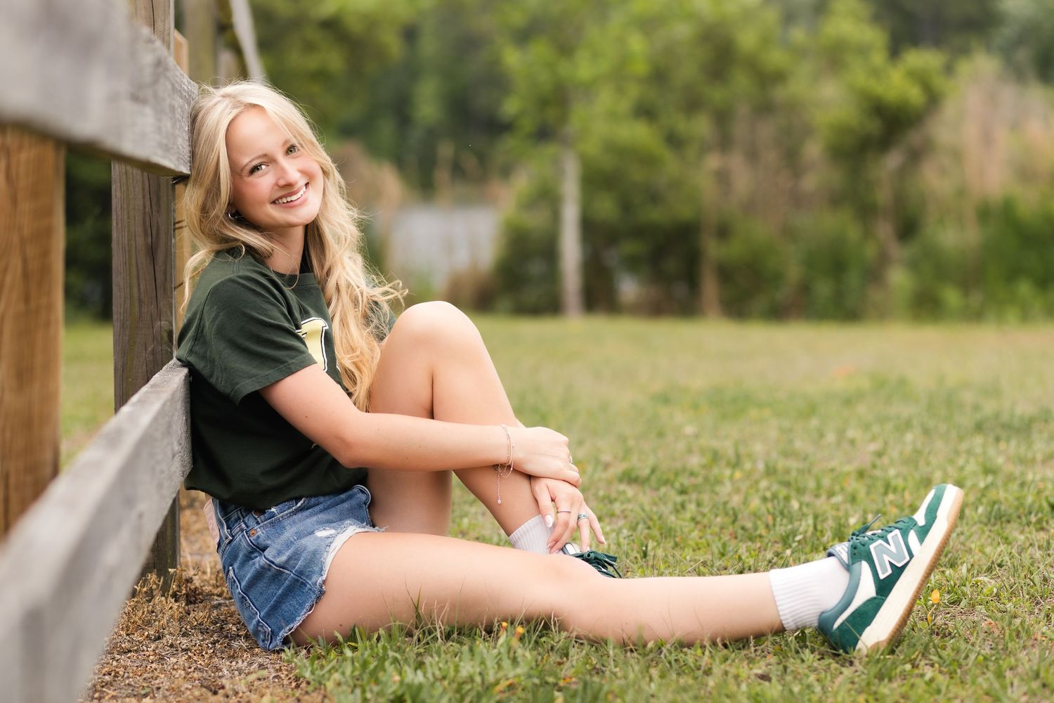 Smiling teenage girl in green t-shirt and denim shorts sitting by a wooden fence outdoors.
